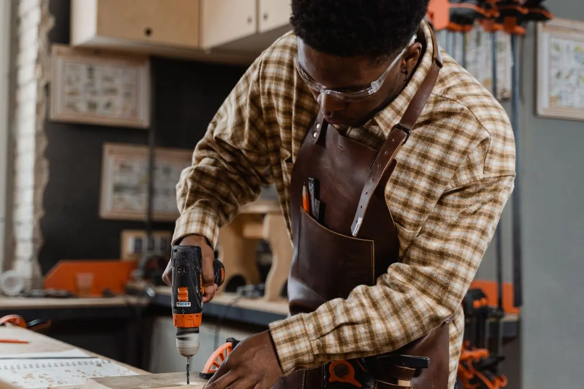 A joiner in goggles and a leather apron drilling at the workshop bench