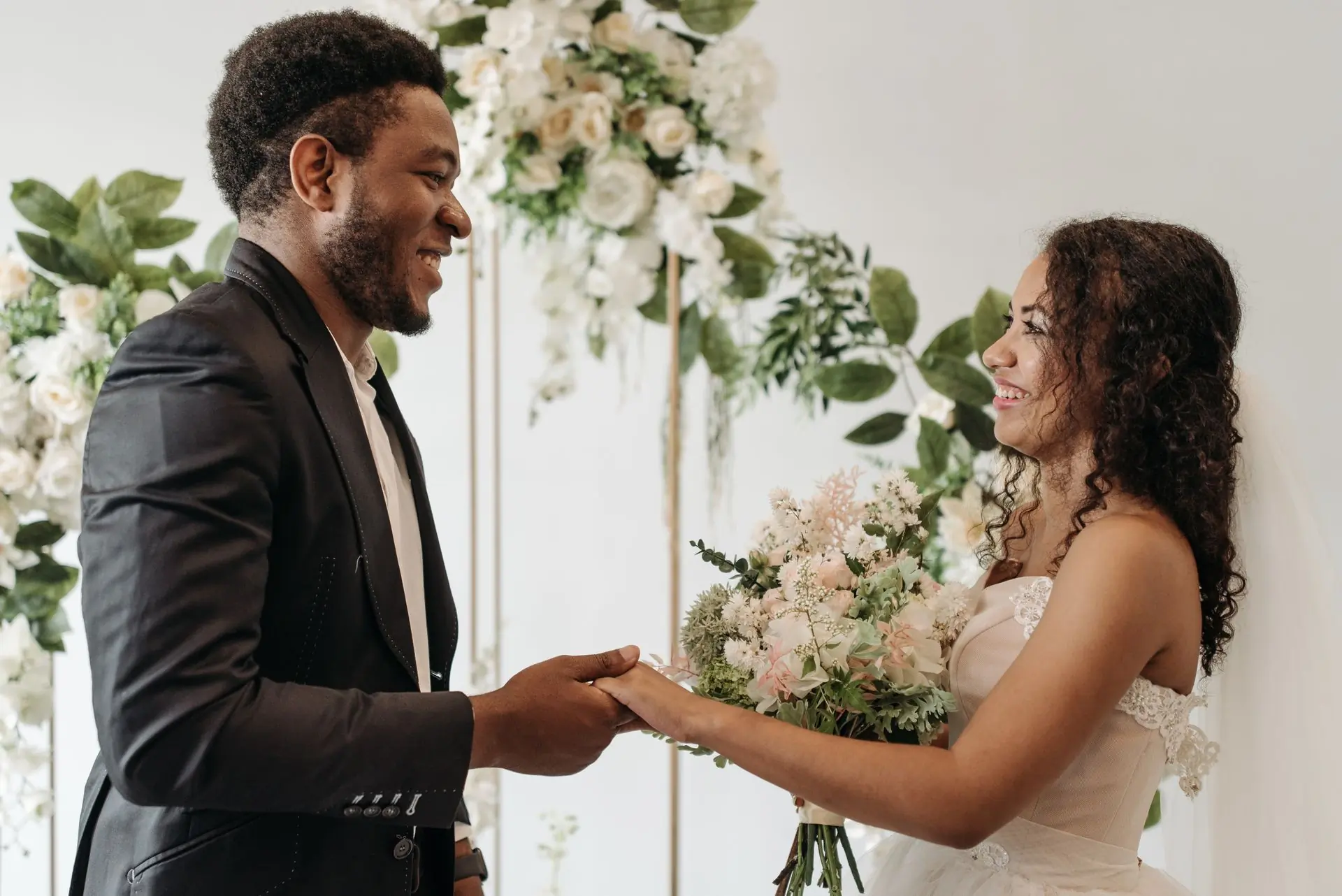 A couple holding hands in front of a pale floral arch on their wedding day