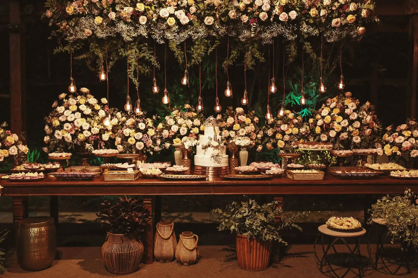 A long reception table lit by hanging bulbs under a canopy of white flowers