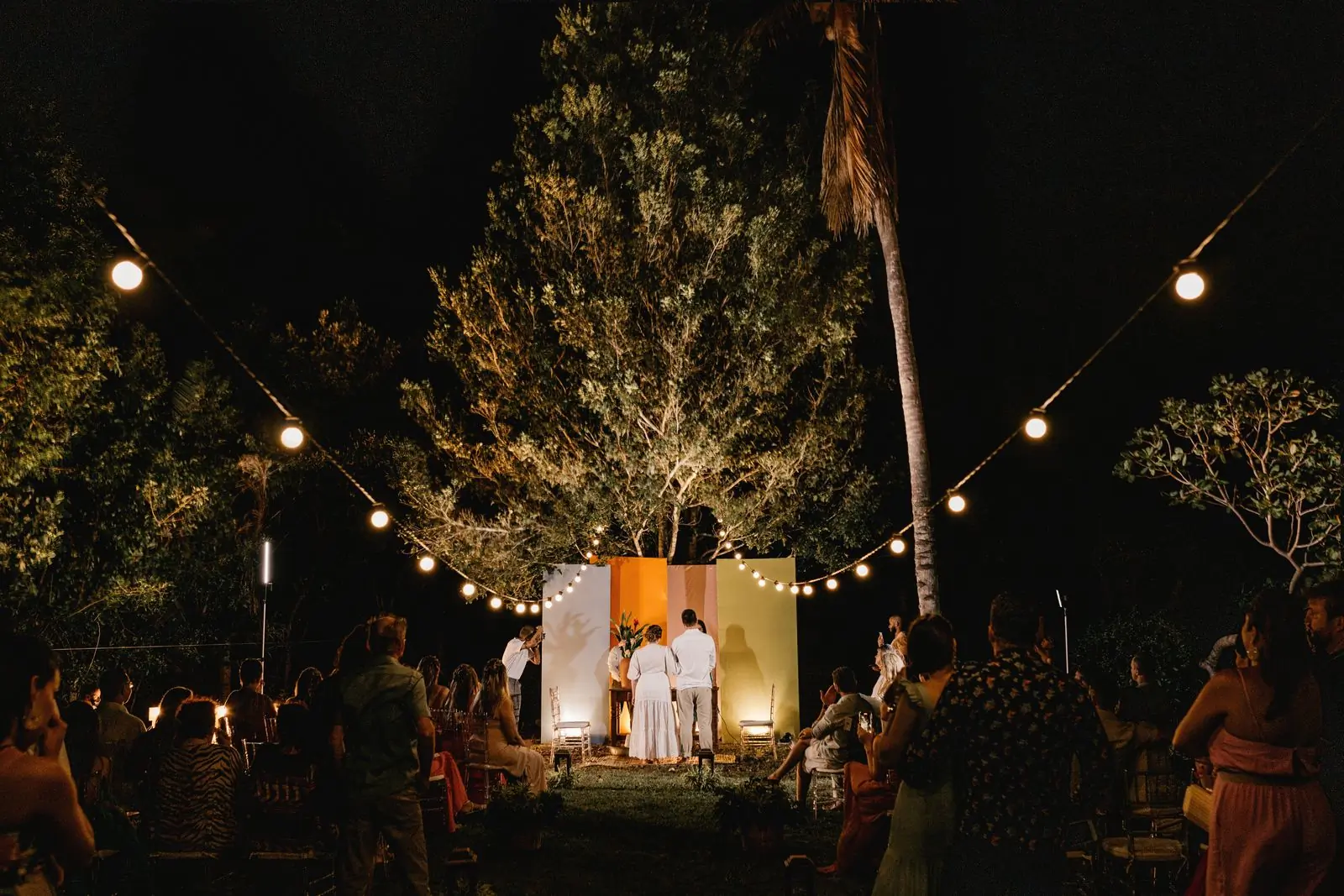 An outdoor evening ceremony lit by strings of bulbs strung between trees