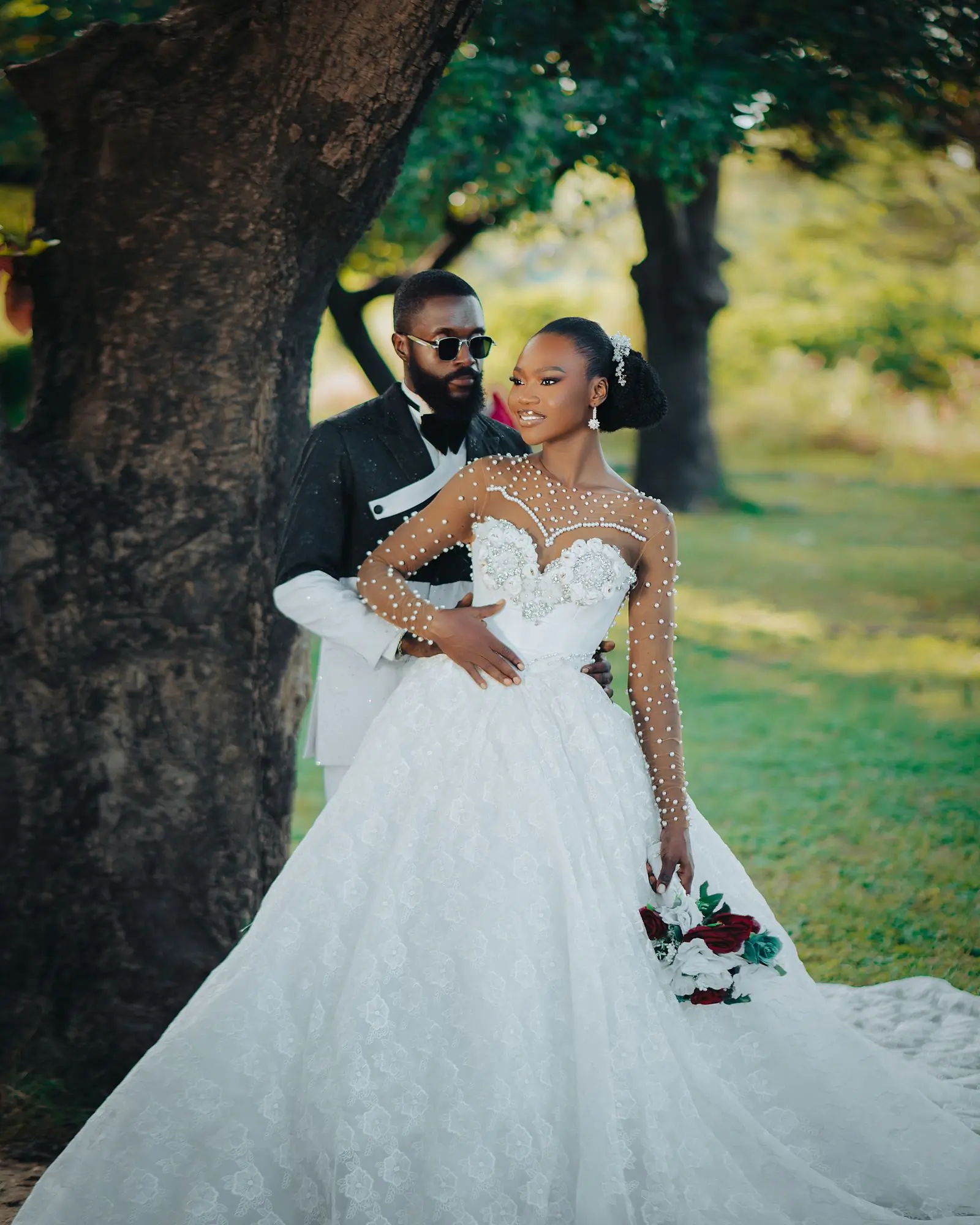 A couple in a beaded gown and dark suit standing under trees in late afternoon light