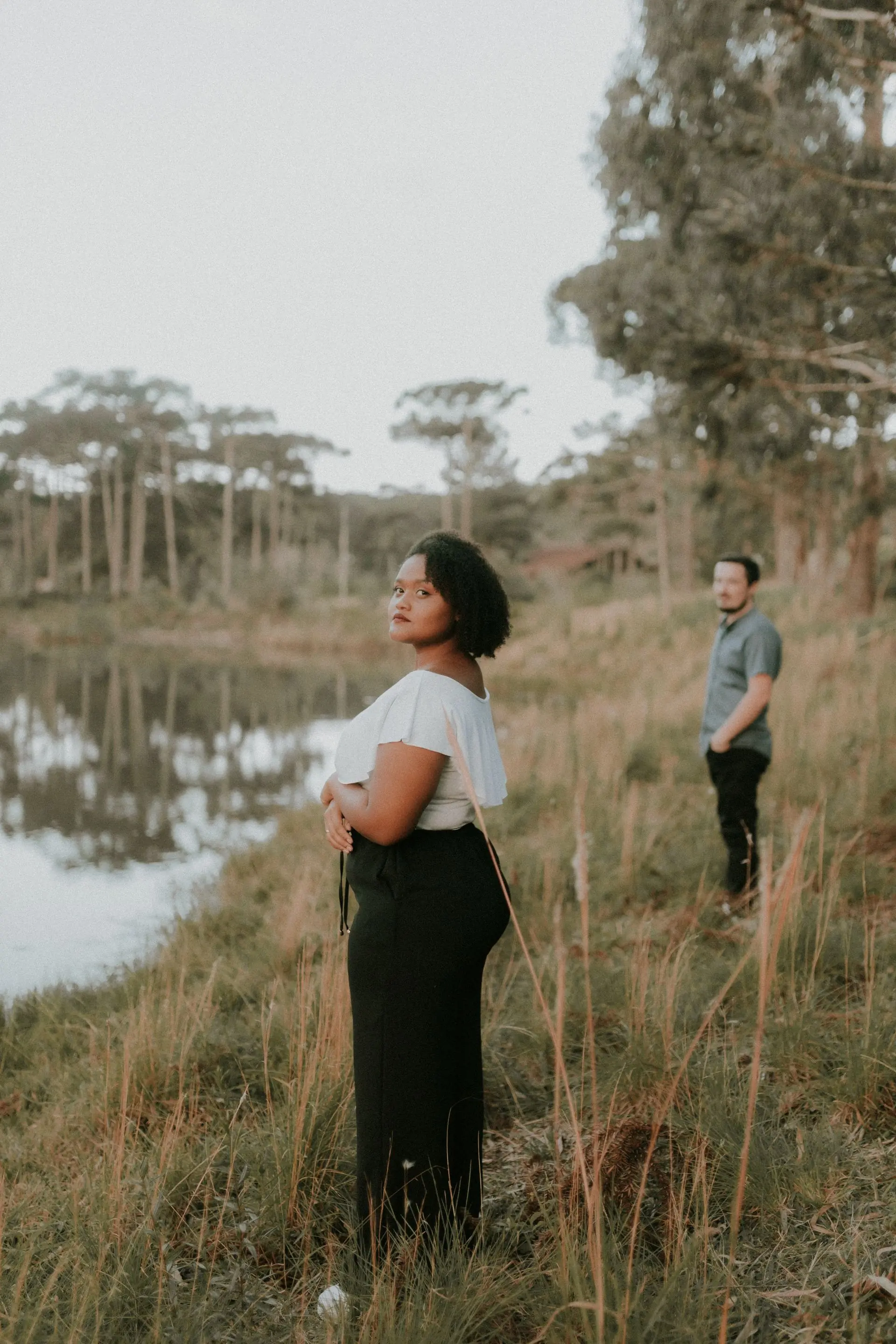 A couple standing apart at the edge of a lake, photographed among tall trees