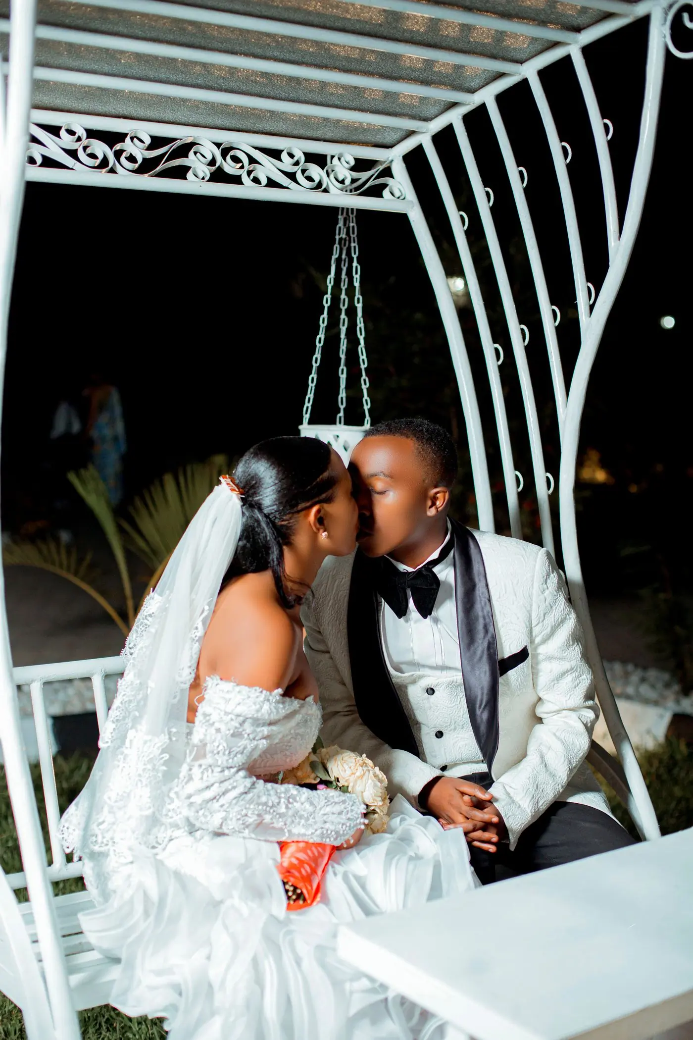 A bride and groom kissing on a white swing seat at dusk