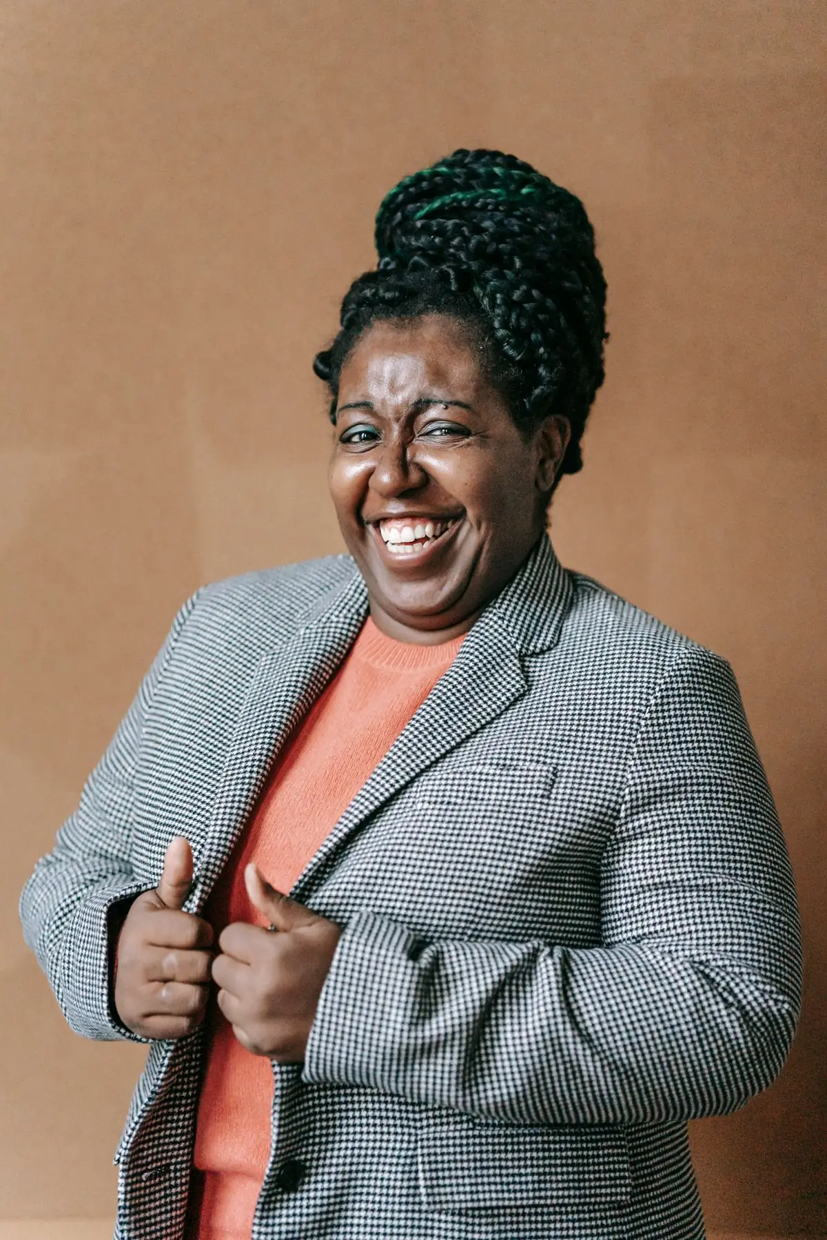 A Black woman with braids pinned up in a high bun, laughing in a grey houndstooth blazer over a coral jumper, against a plain warm tan wall