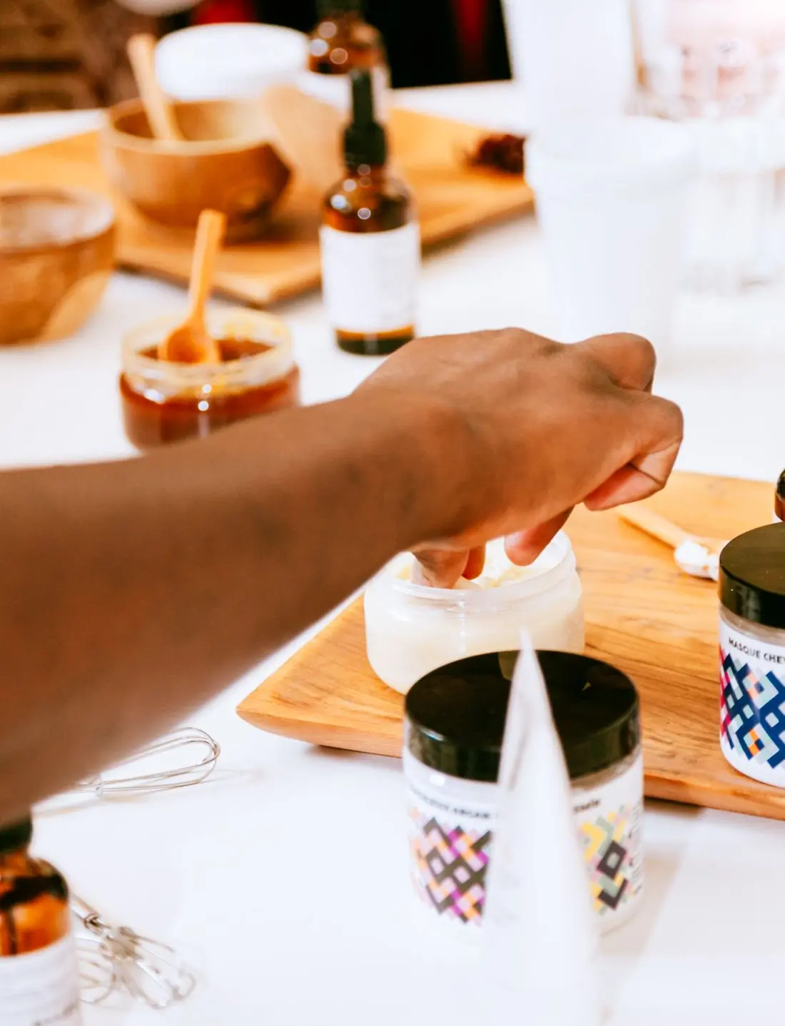 A brown hand scooping soft white balm from an open tub on a wooden board, amber dropper bottles, wooden bowls and a small whisk on the white bench around it