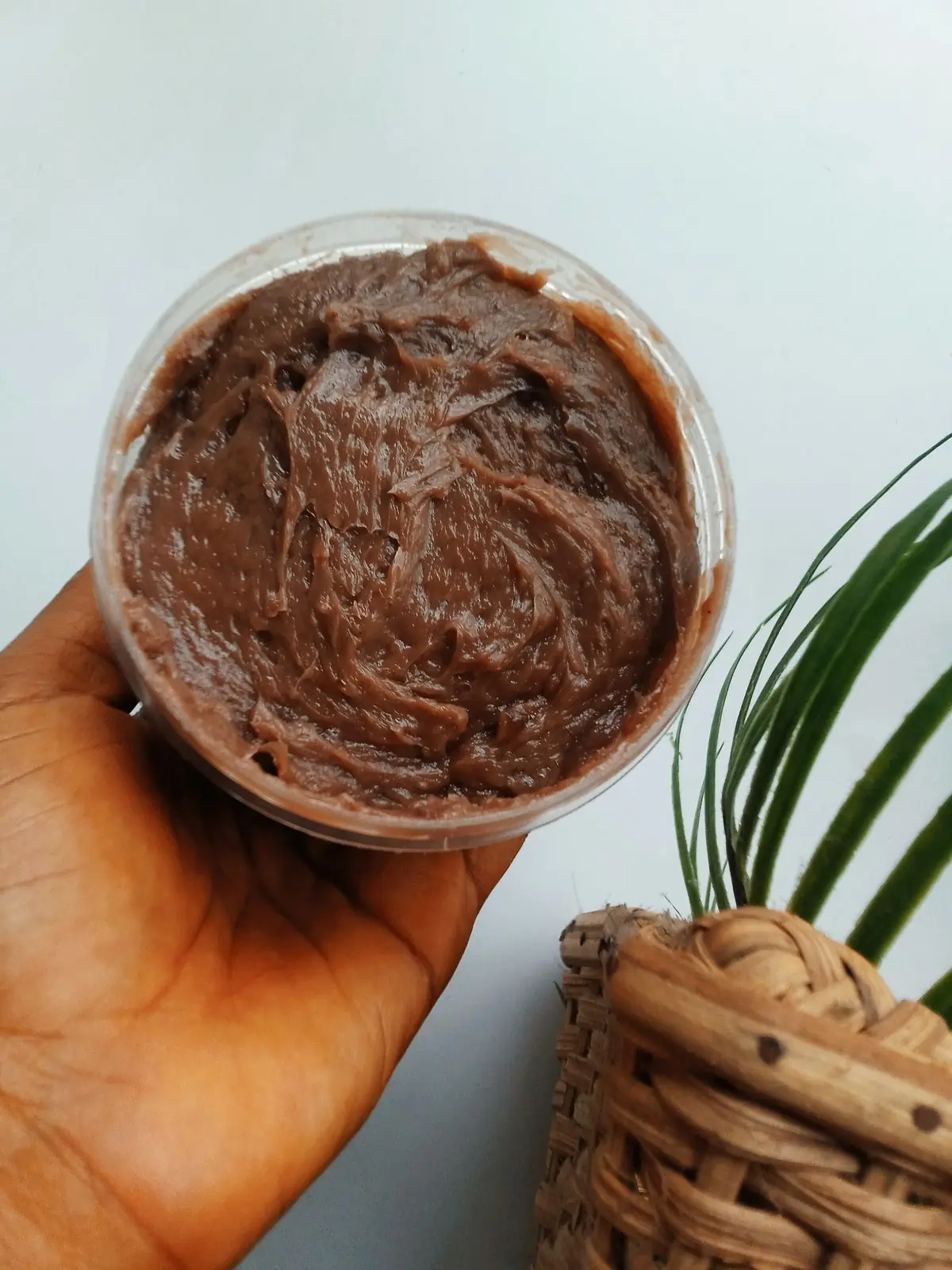 A brown hand holding up a clear tub of whipped chocolate brown body butter beside a woven basket and a palm frond