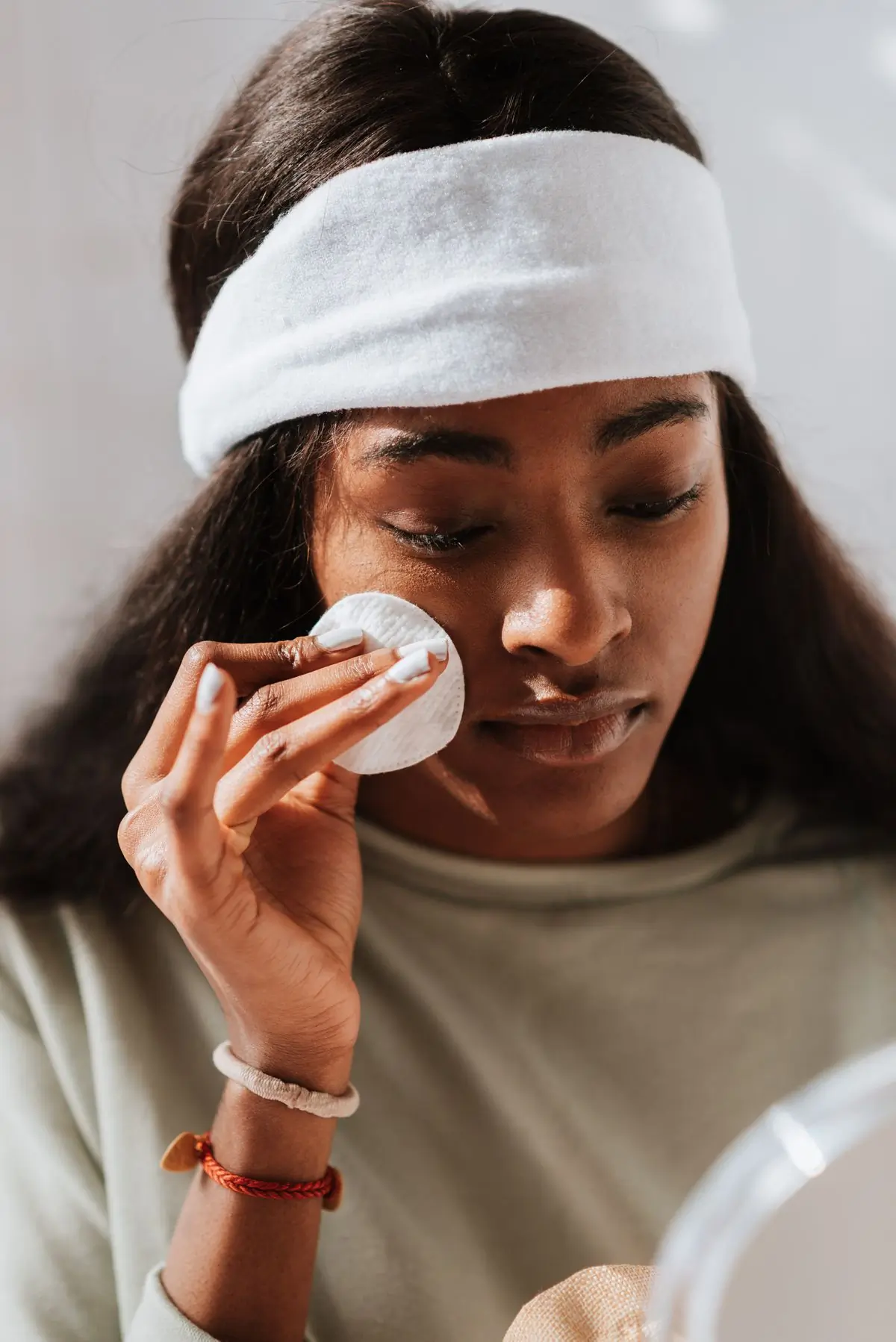 A Black woman in a white towelling headband wiping her cheek with a round cotton pad, eyes lowered against a plain cream wall