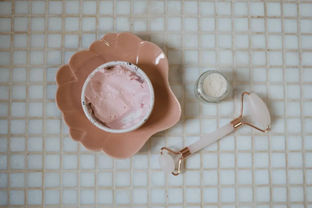 A pot of pink clay mask in a scalloped peach dish beside a rose quartz roller, laid on pale mosaic tile
