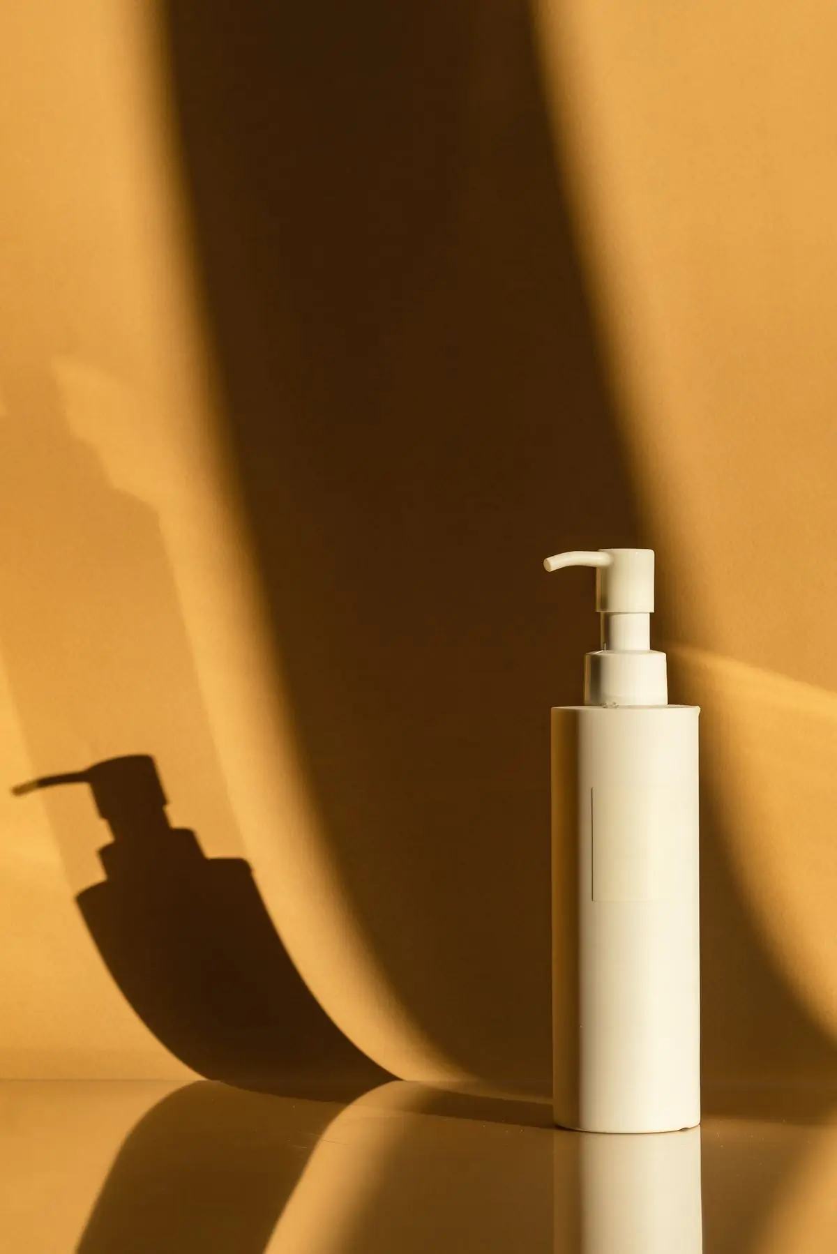 An unlabelled white pump bottle standing in hard sunlight on a warm amber ground, throwing a long shadow