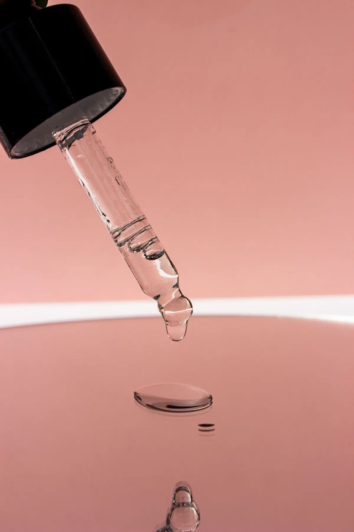 A black capped glass dropper held tip down over a glossy ledge, one clear bead about to fall, against a dusty rose backdrop