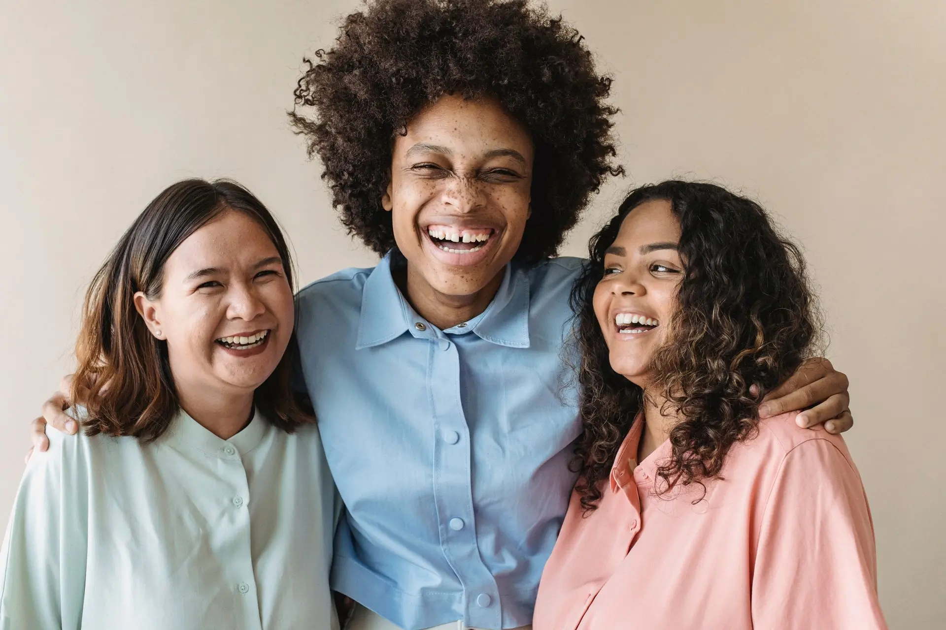 Three friends laughing with their arms round each other, a Black woman with an afro in the middle, in mint, pale blue and peach shirts against a warm beige wall