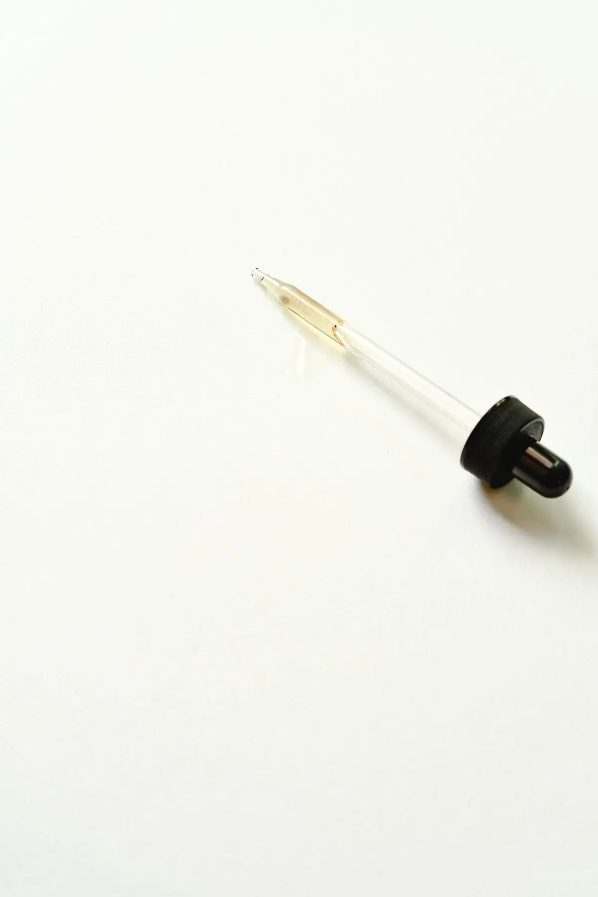 A glass dropper half full of amber serum lying on a bare ivory surface with empty space all around it