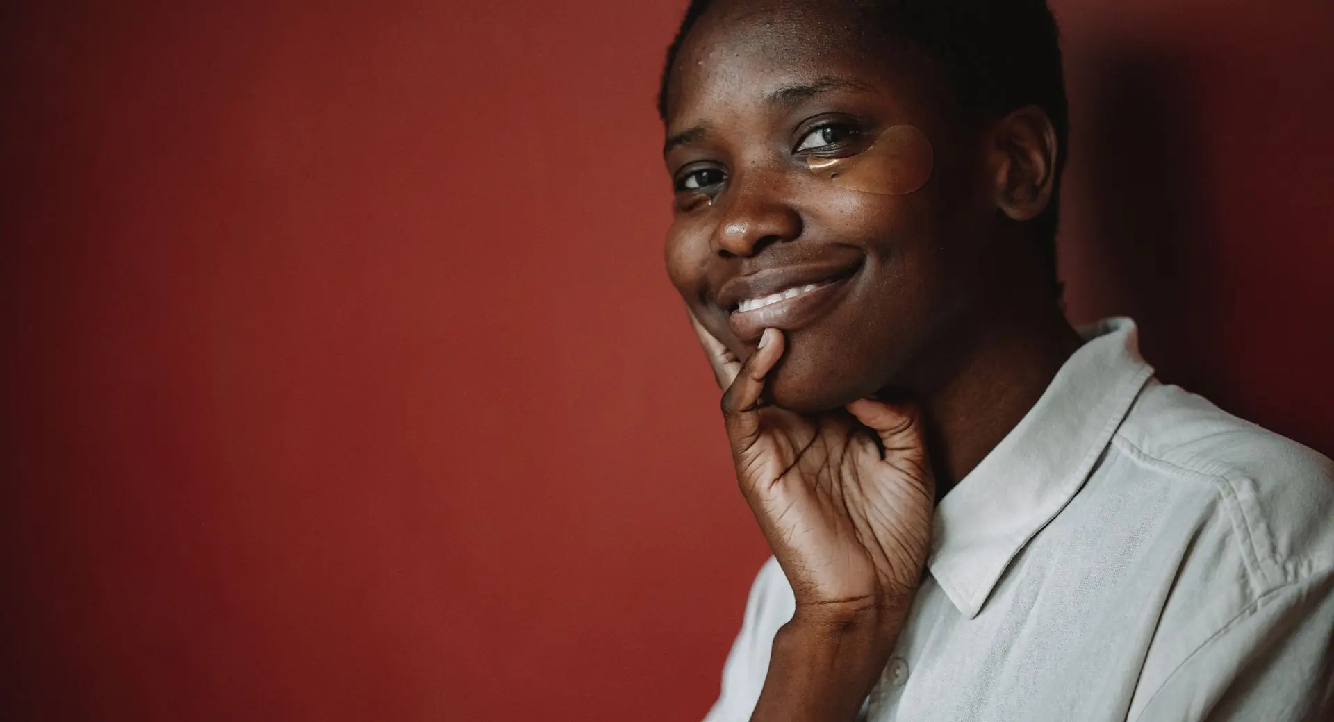 A Black woman in a cream shirt resting her chin on her hand and smiling, a gel patch under one eye, against a plain deep terracotta wall