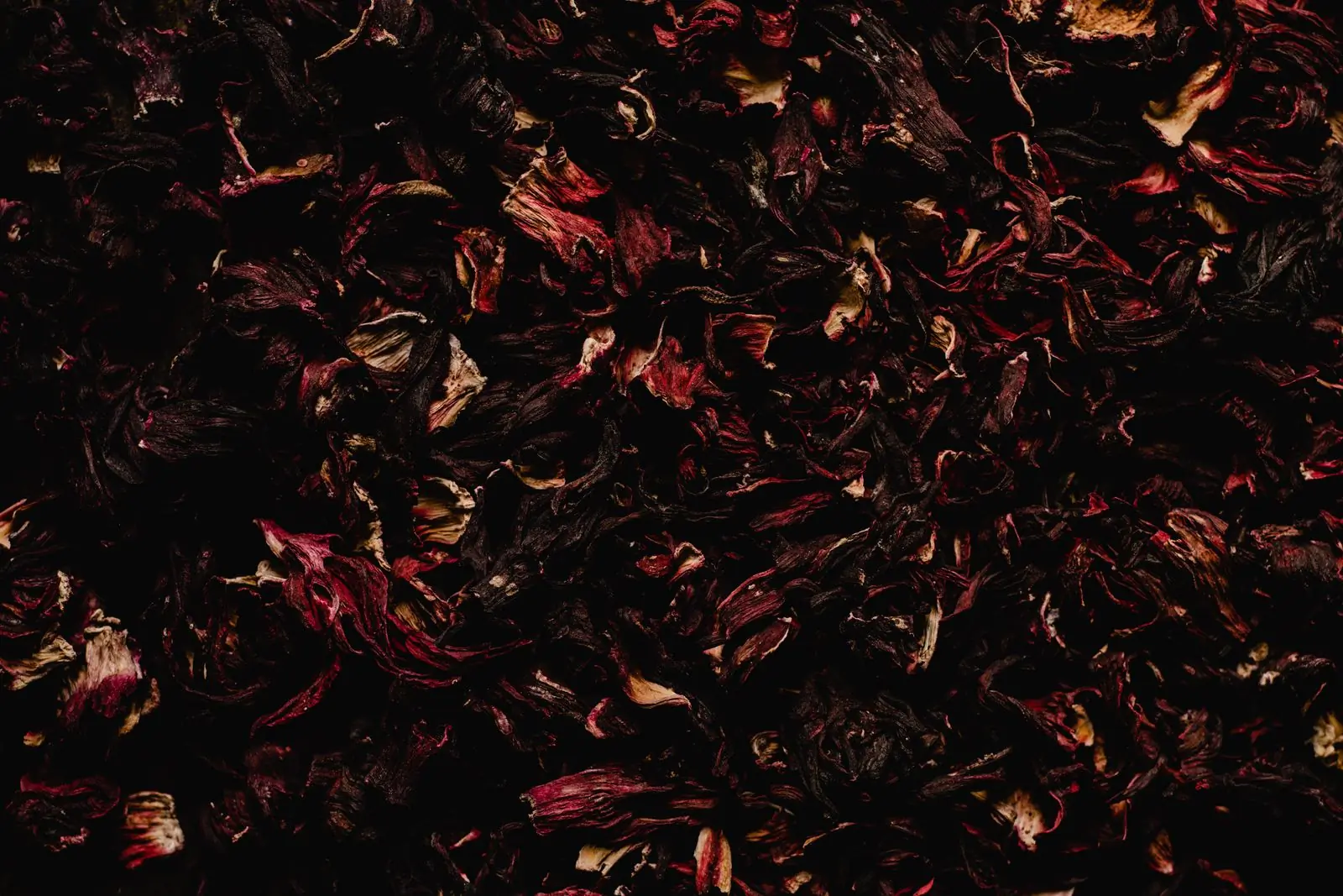 A dense bed of dried hibiscus petals filling the frame, deep burgundy shading to rust where the light catches the curled edges