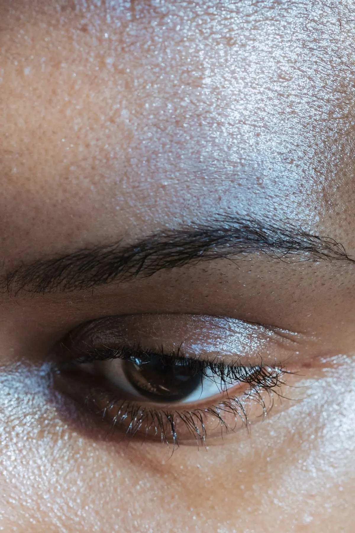 An extreme close-up of a Black woman's eye and brow, brown skin catching a dewy highlight across the lid and temple