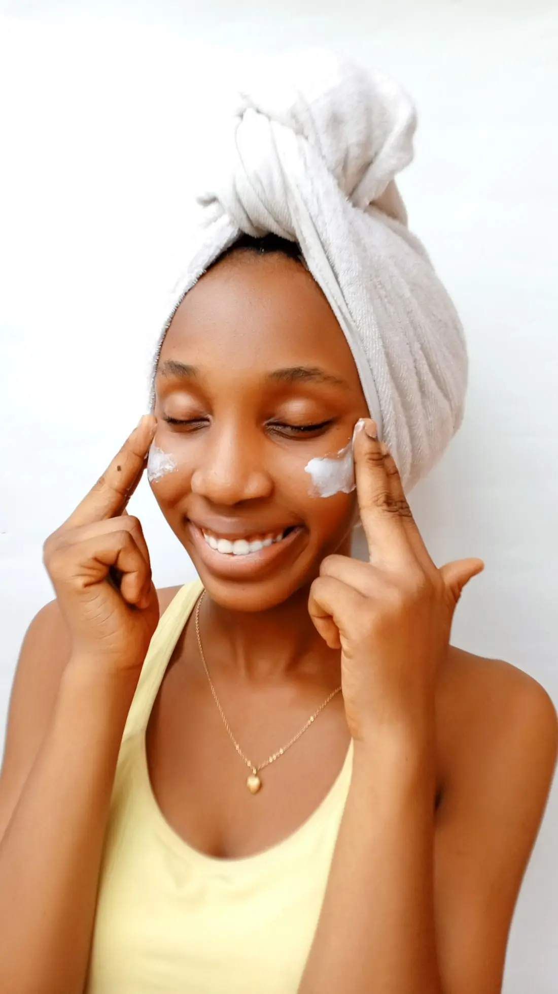 A Black woman with a towel wrapped round her hair, eyes closed and smiling, pressing a dab of white cream into each cheek