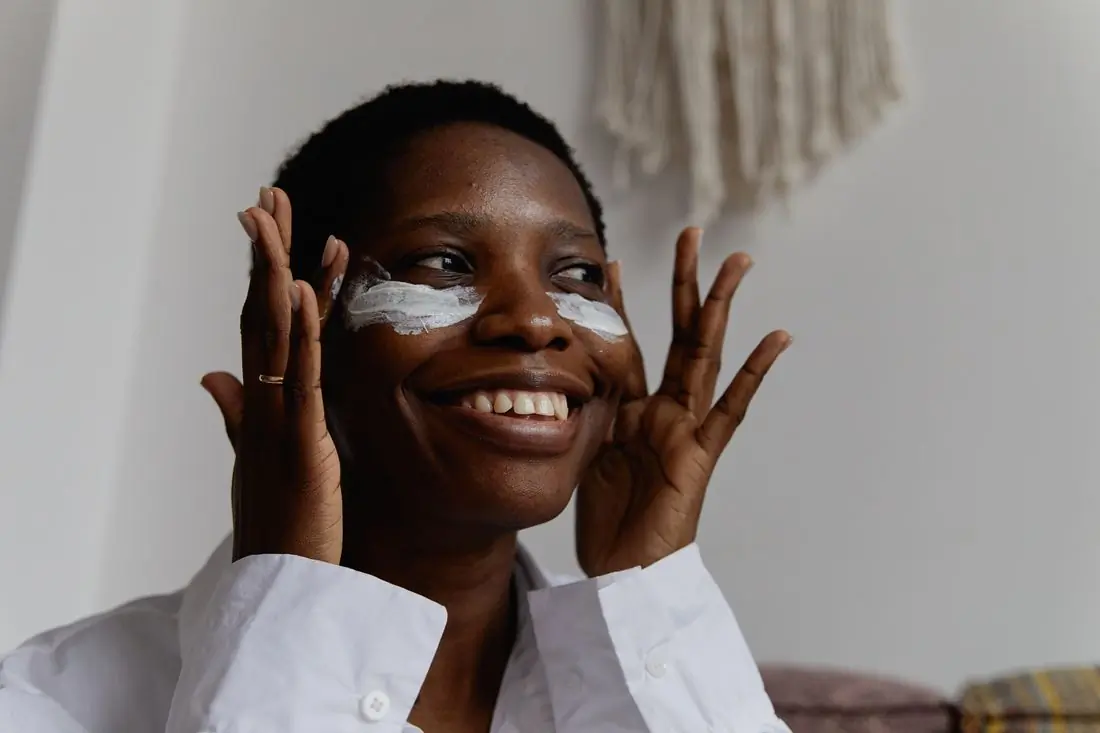 A Black woman in a white shirt laughing with stripes of white cream under both eyes, hands raised beside her face