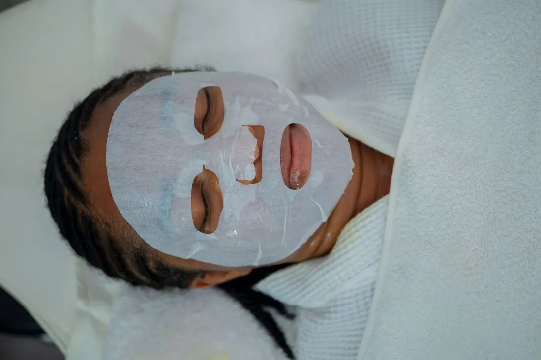 A Black woman lying back with a wet white sheet mask smoothed over her face, wrapped in white towels