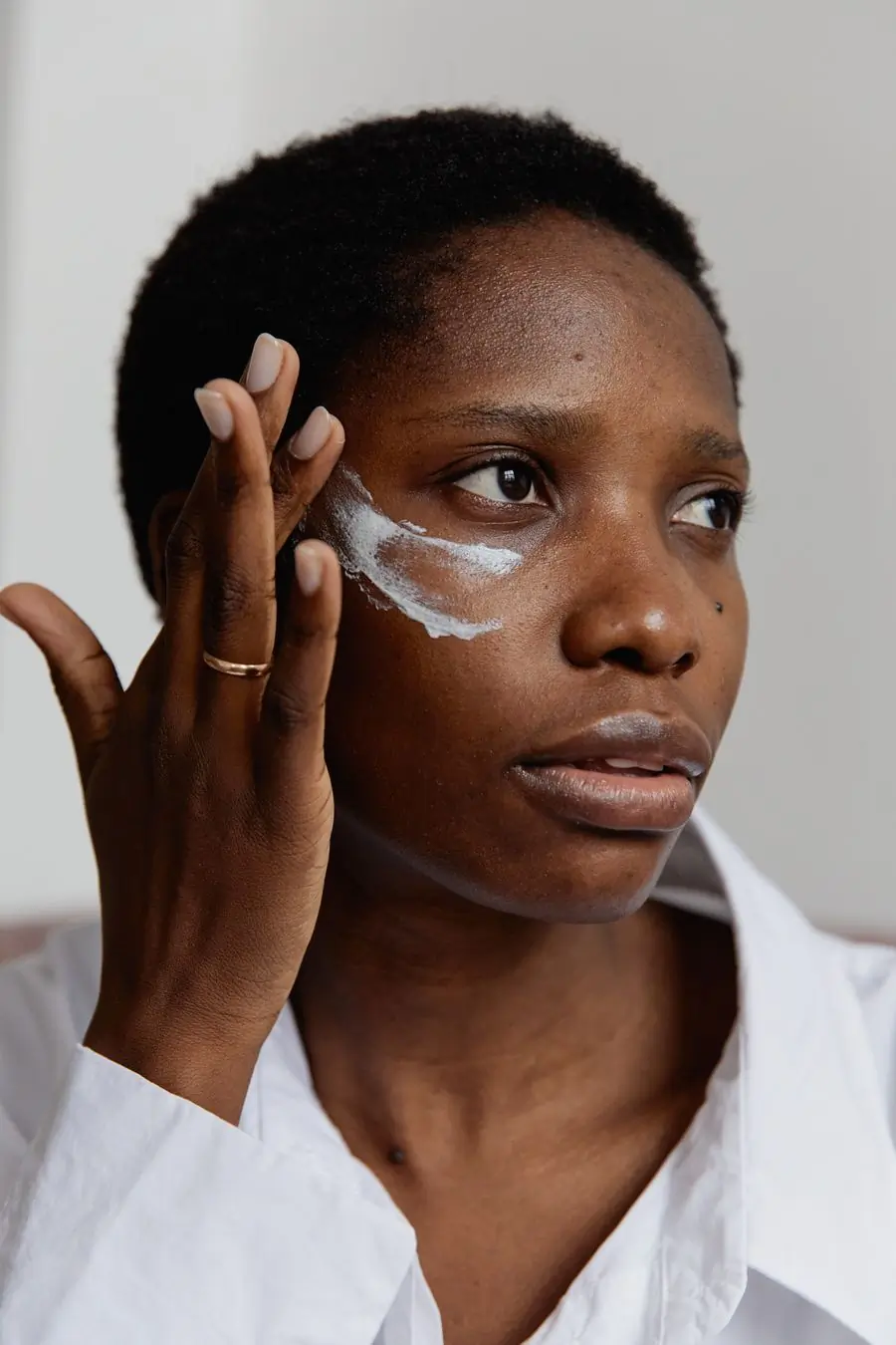 A Black woman in a white shirt pressing white cream into her cheek with two fingertips, plain pale wall behind