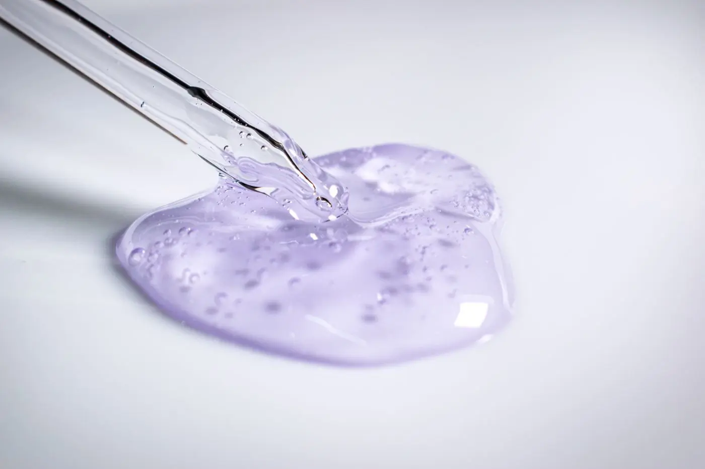 A glass pipette releasing a pool of clear lilac gel, small bubbles caught in it, on a bright white ground