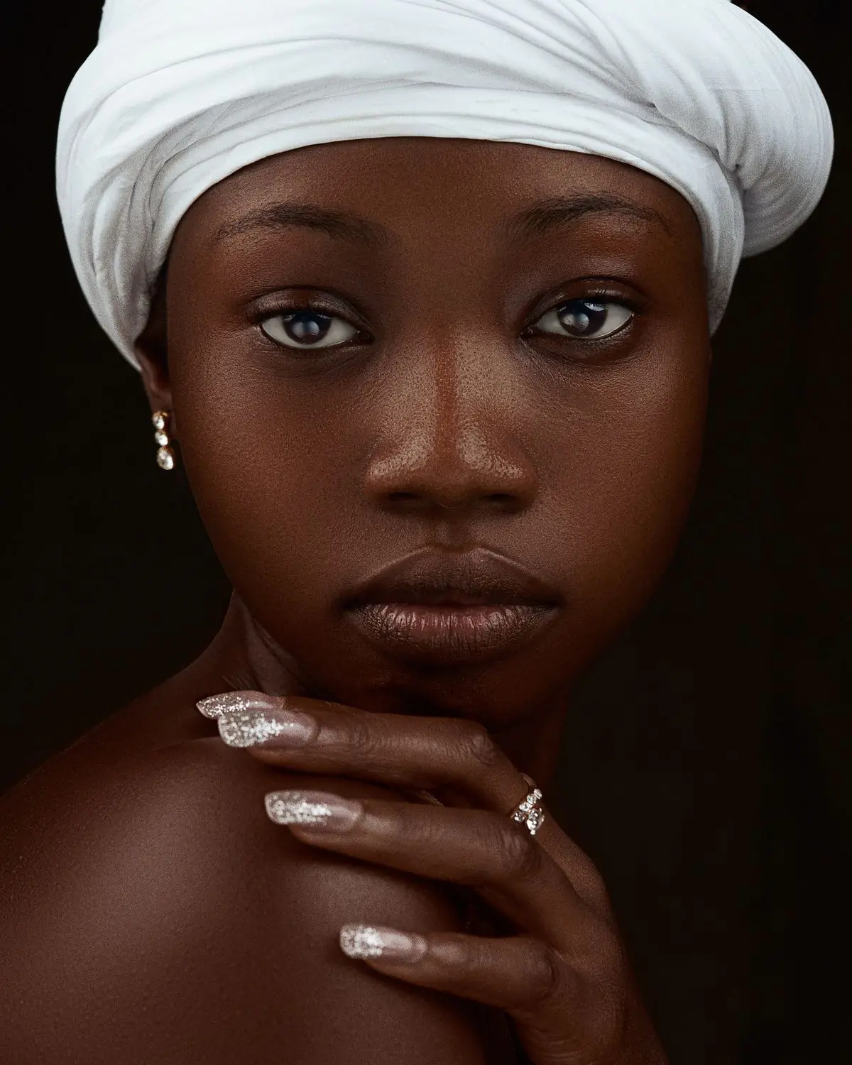 A Black woman in a white head wrap resting her chin on her hand, looking to camera against a near black ground