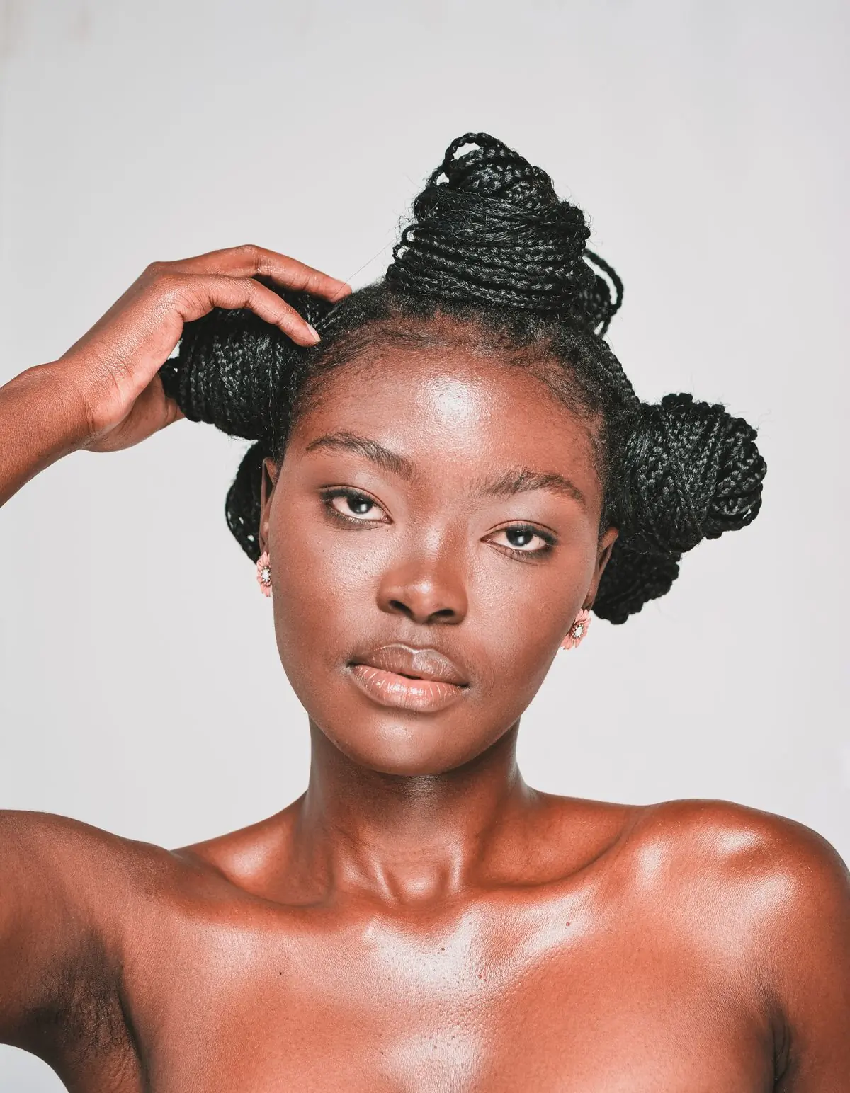 A Black woman with her braids pinned into buns, one hand lifted to her hair, bare shoulders against a pale studio ground