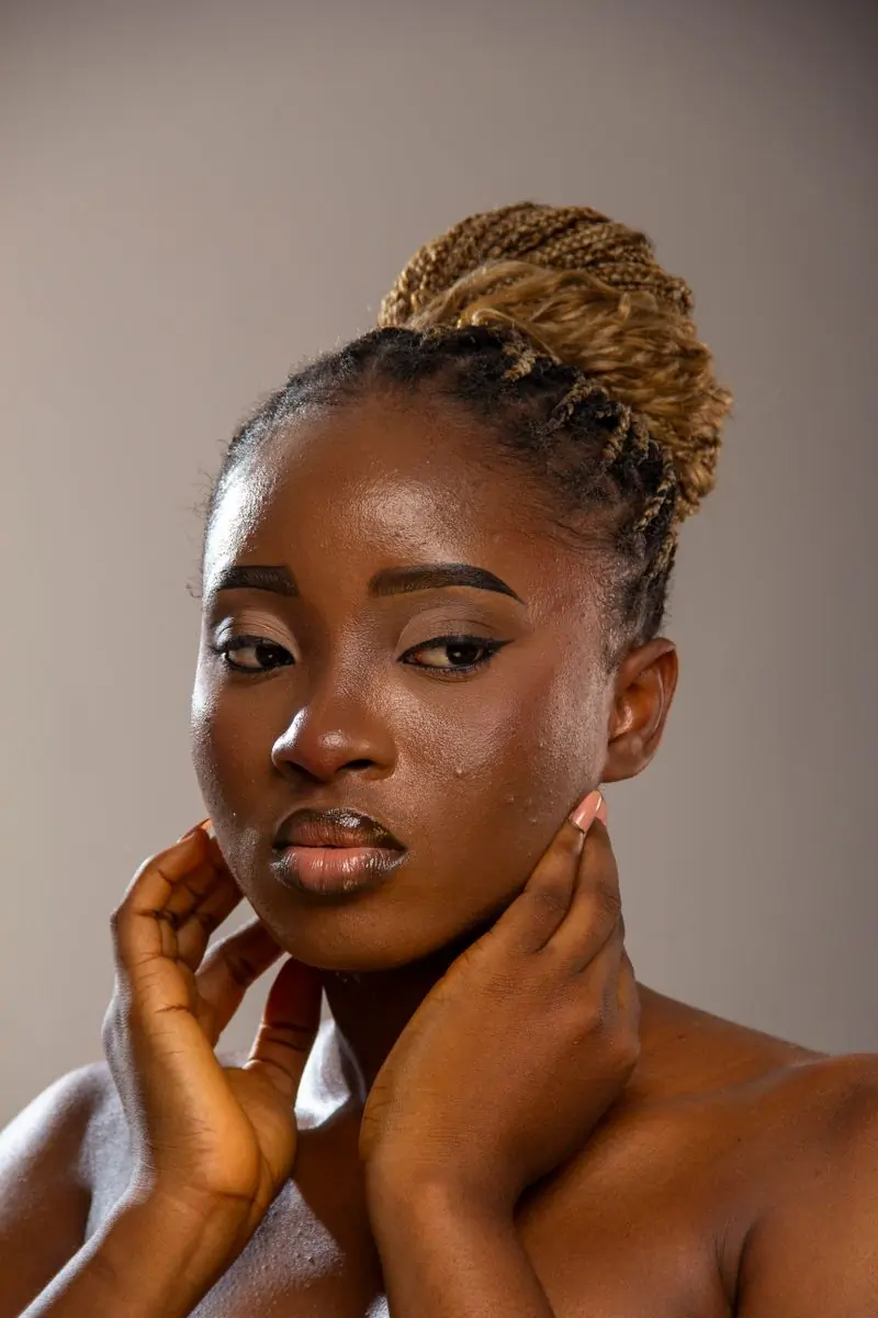 A Black woman with honey blonde braids pinned into a high bun, both hands raised to her neck, against a plain warm grey ground