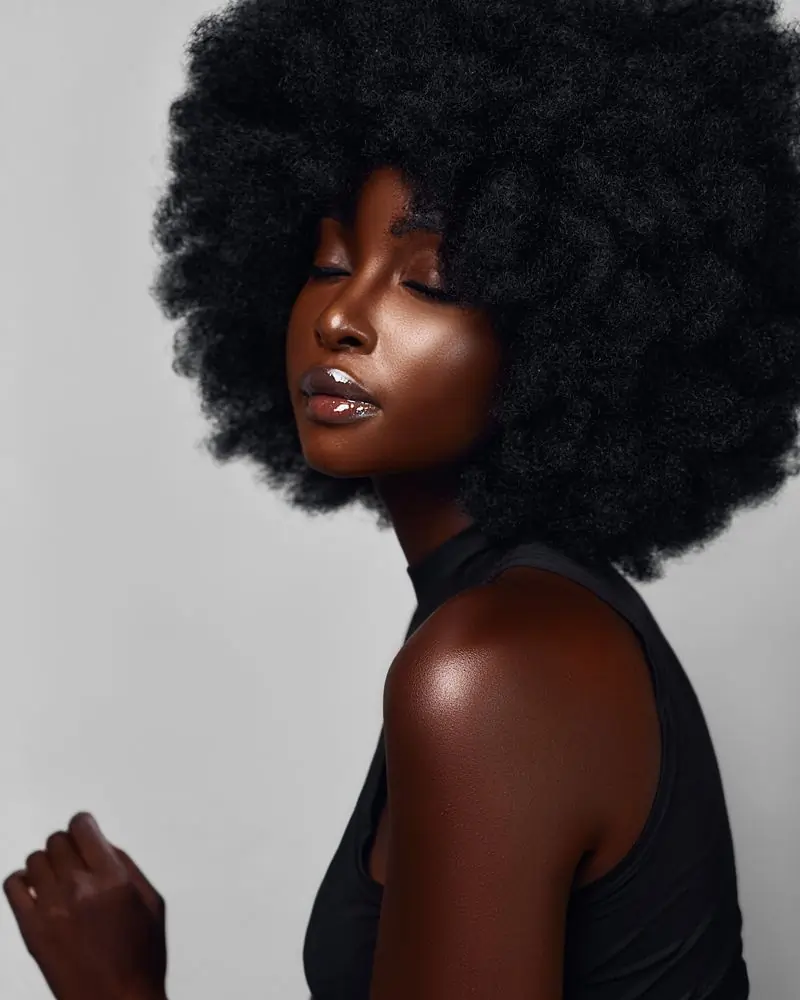 A Black woman with a large round afro in profile, eyes closed and chin lifted, against a plain pale grey ground