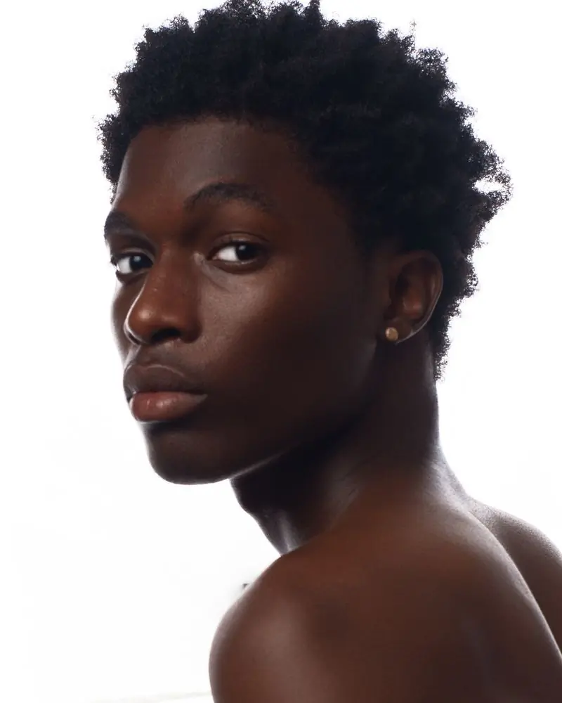 A young Black man with a short afro and a small gold stud earring, looking back over his bare shoulder against a plain white ground