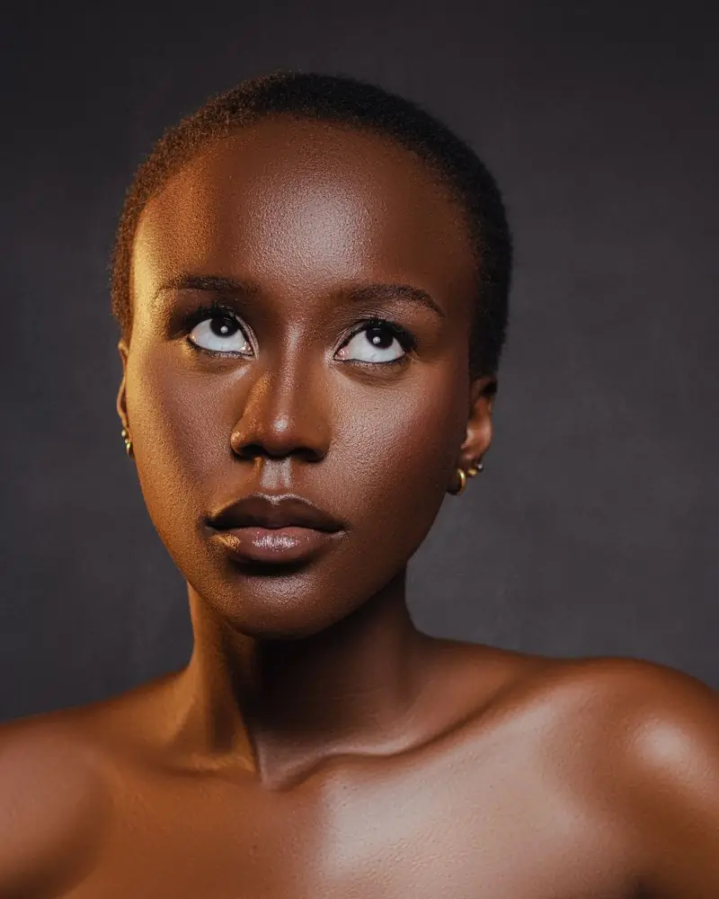 A Black woman with closely cropped hair and small gold earrings looking up and away, bare shoulders against a dark charcoal ground