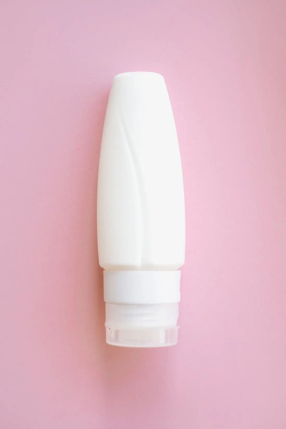 A plain white unlabelled lotion bottle lying cap downwards on a flat rose pink paper ground