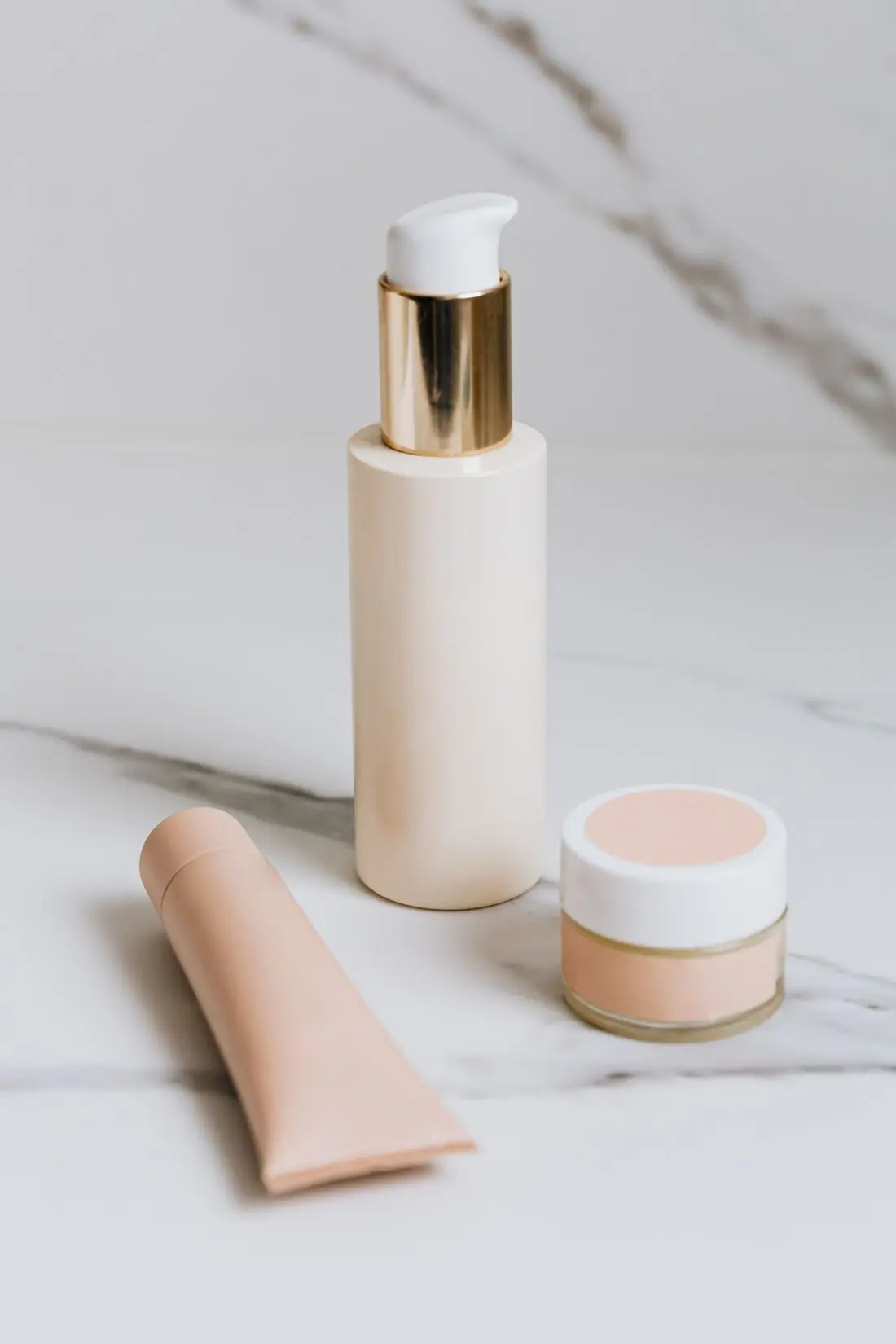 A cream pump bottle with a gold collar standing on white marble, a peach squeeze tube lying beside it and a small glass pot of peach cream alongside