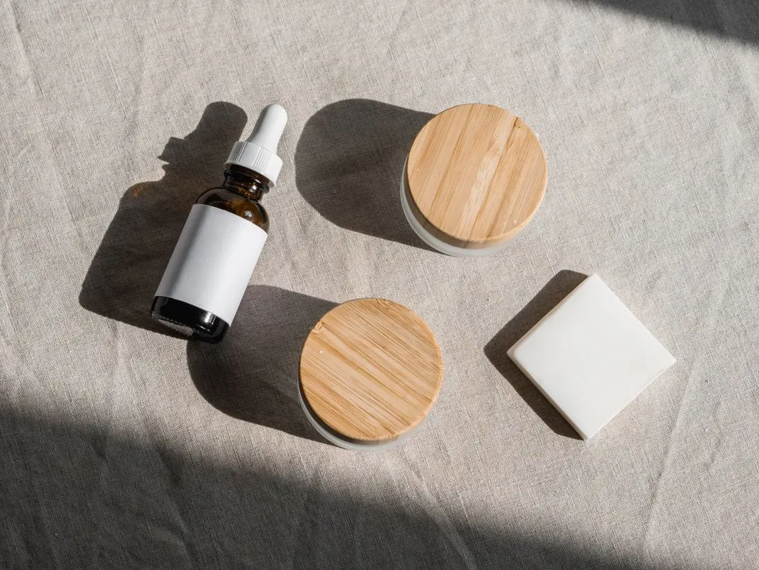An amber dropper bottle with a blank label, two bamboo lidded jars and a soap bar on oatmeal linen in raking sunlight