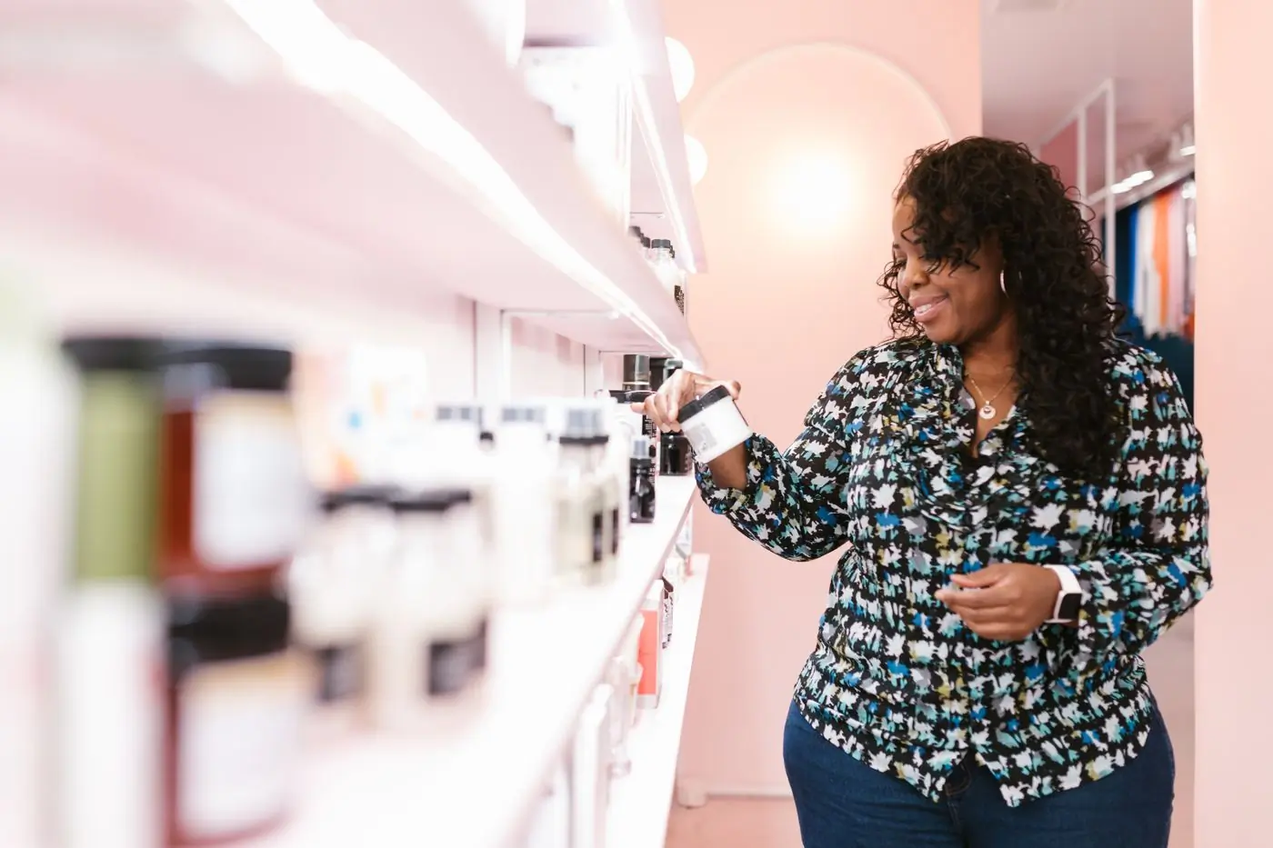 A Black woman in a floral blouse smiling as she lifts a white jar with a black lid from a long white shelf in a pink-walled shop