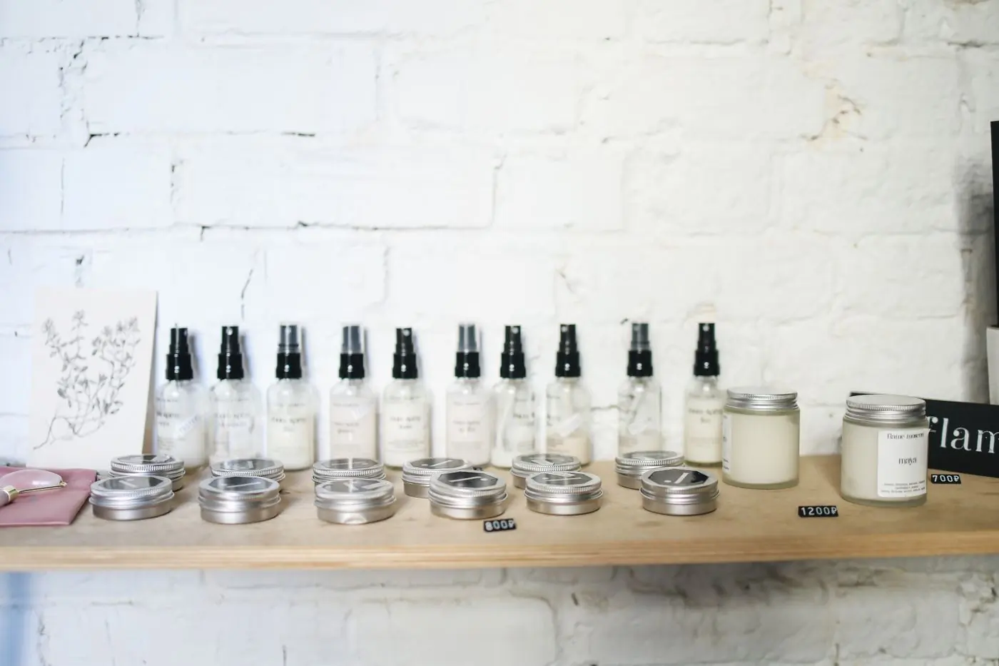 A pine shelf on a whitewashed brick wall holding a row of clear spray bottles with black pump tops, a line of round silver tins in front of them and two candles in glass jars at the end