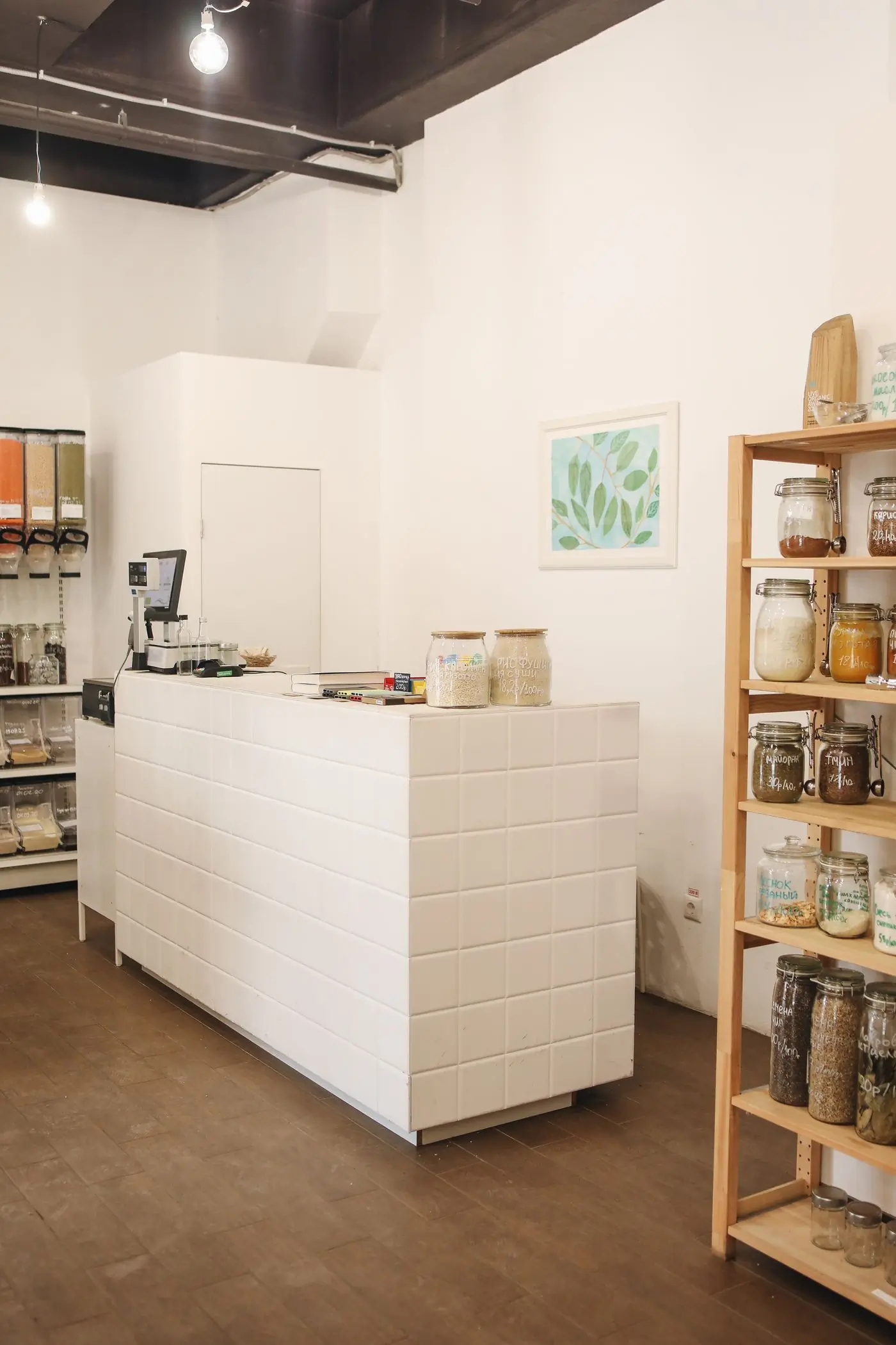 A white tiled service counter with a till and a set of scales on it, a framed leaf print on the wall behind and a tall pine shelf of hand-labelled clamp-top jars beside it