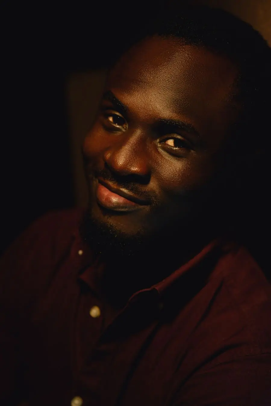A smiling man in a dark red shirt photographed in warm low light