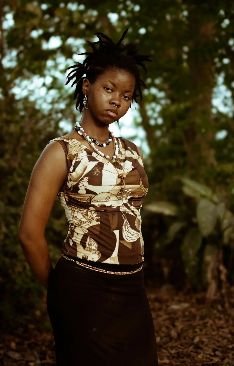 A woman in a patterned top standing among trees, looking at the camera