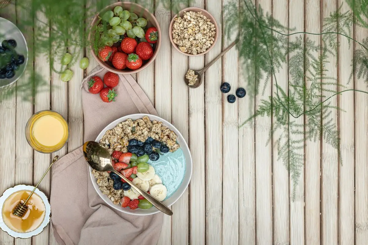 Two breakfast bowls of fruit, granola and honey on a pale wooden table