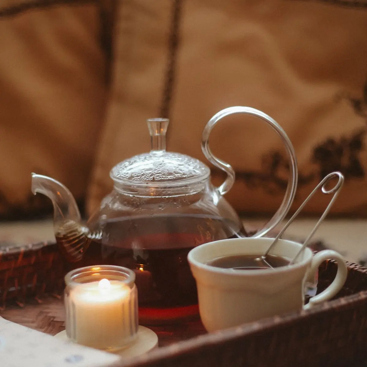 A glass teapot and cup beside a lit candle on a warm wooden table