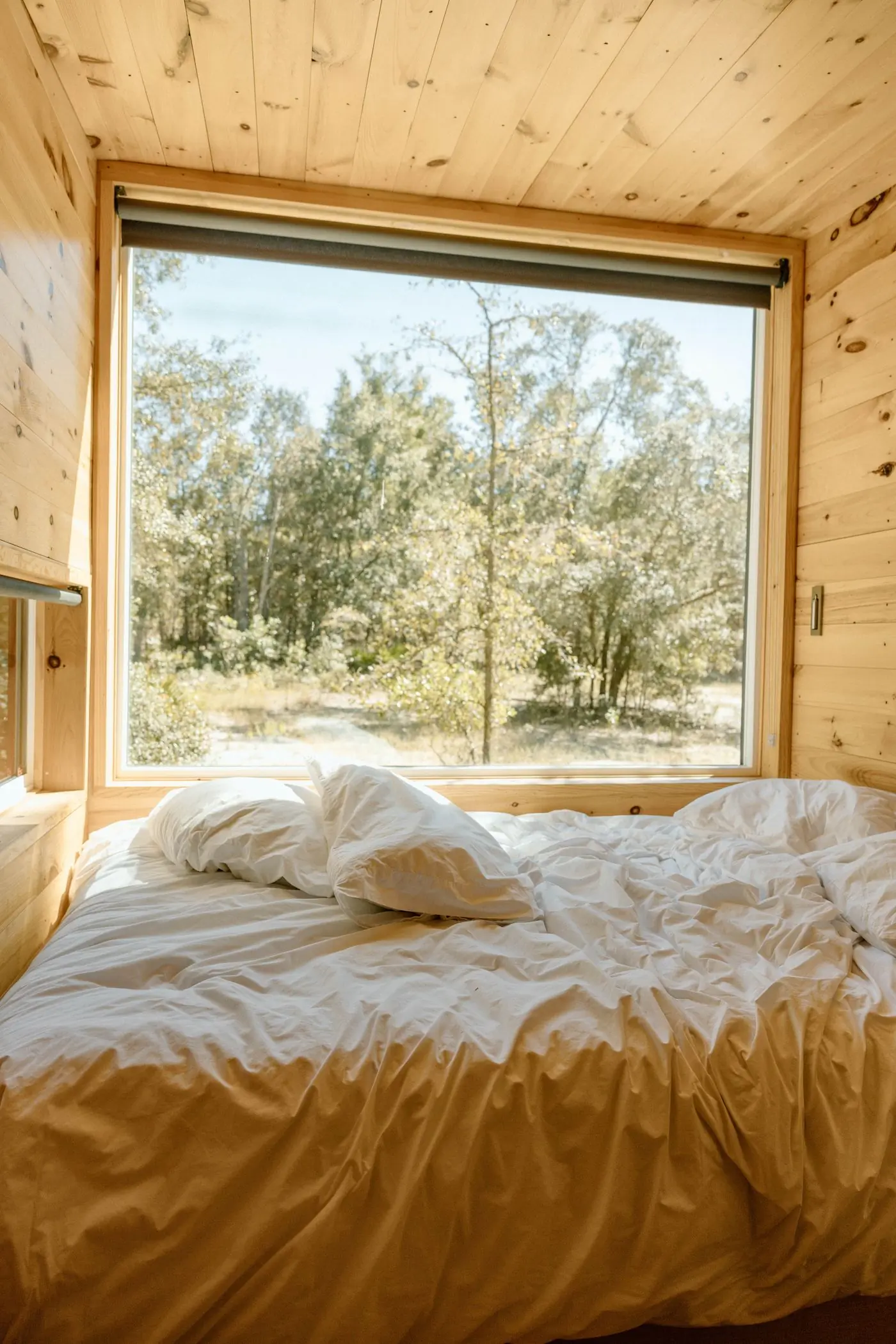 A made bed in a timber room with a wide window onto the forest