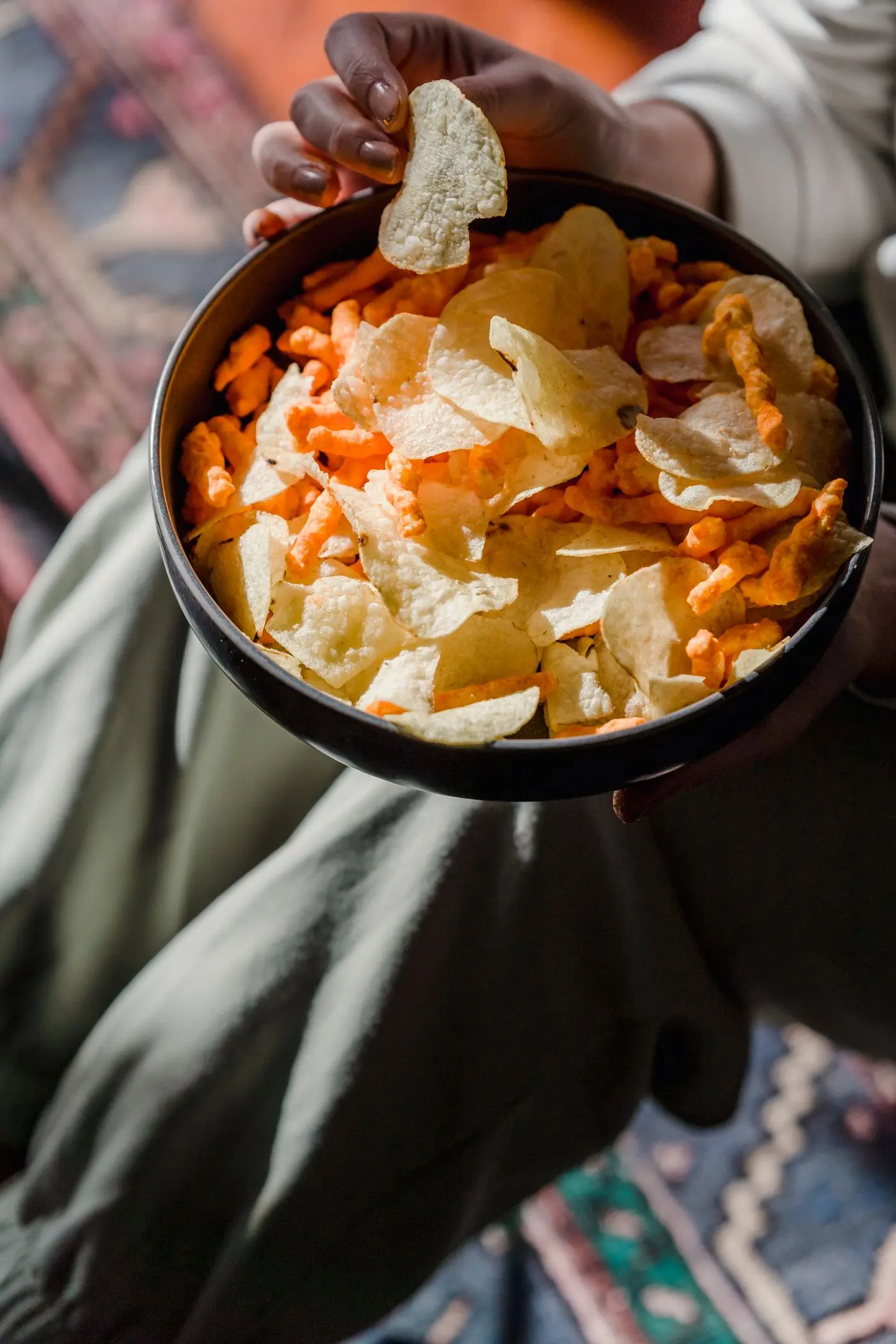 A hand lifting a chip from a bowl resting on a lap, warm rug beneath