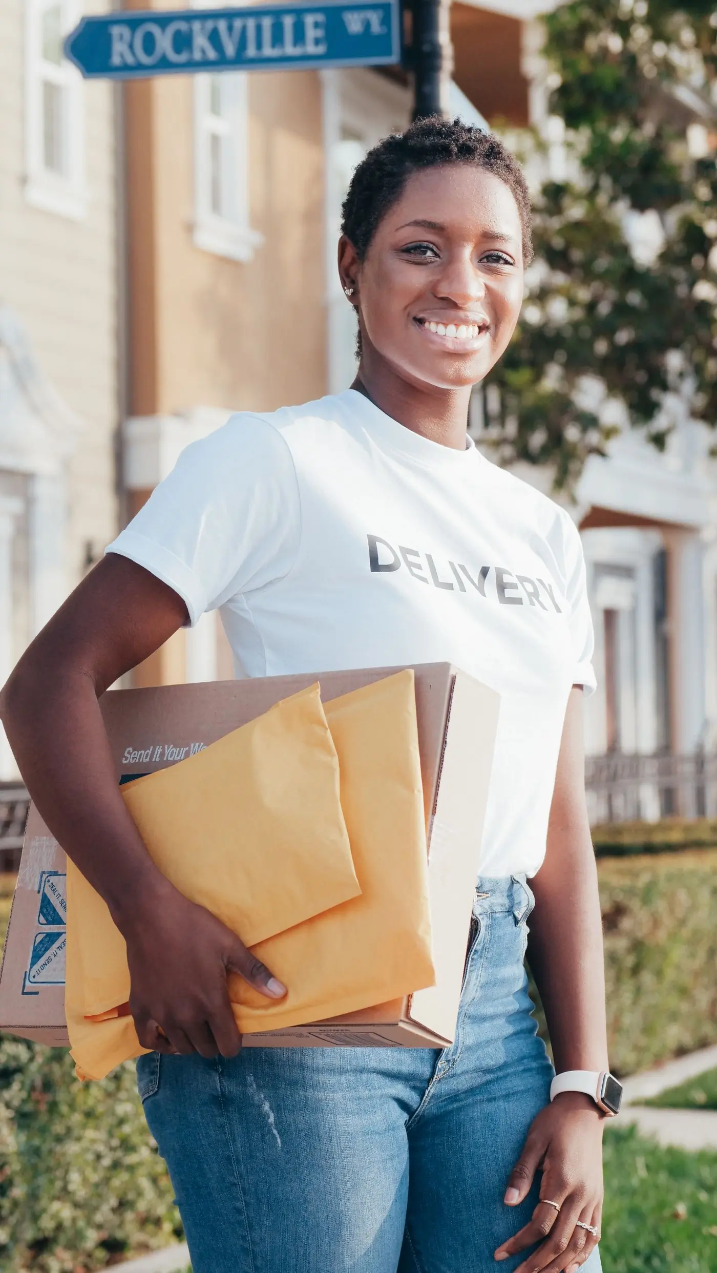 A smiling delivery rider carrying a kraft box and yellow mailers down a sunny street
