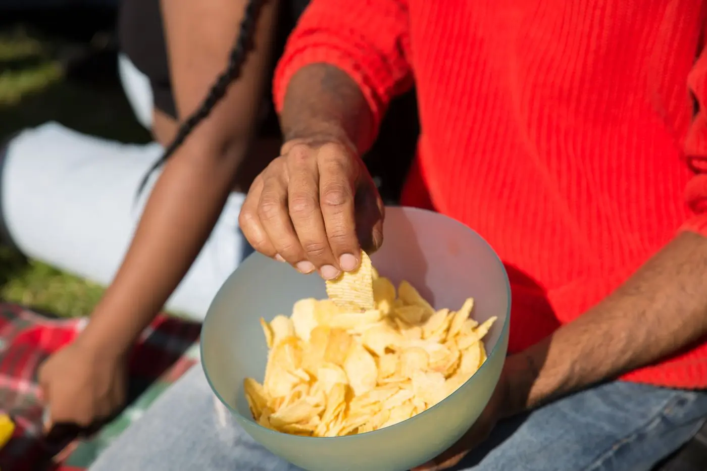A person in a red sweater reaching into a bowl of chips outdoors