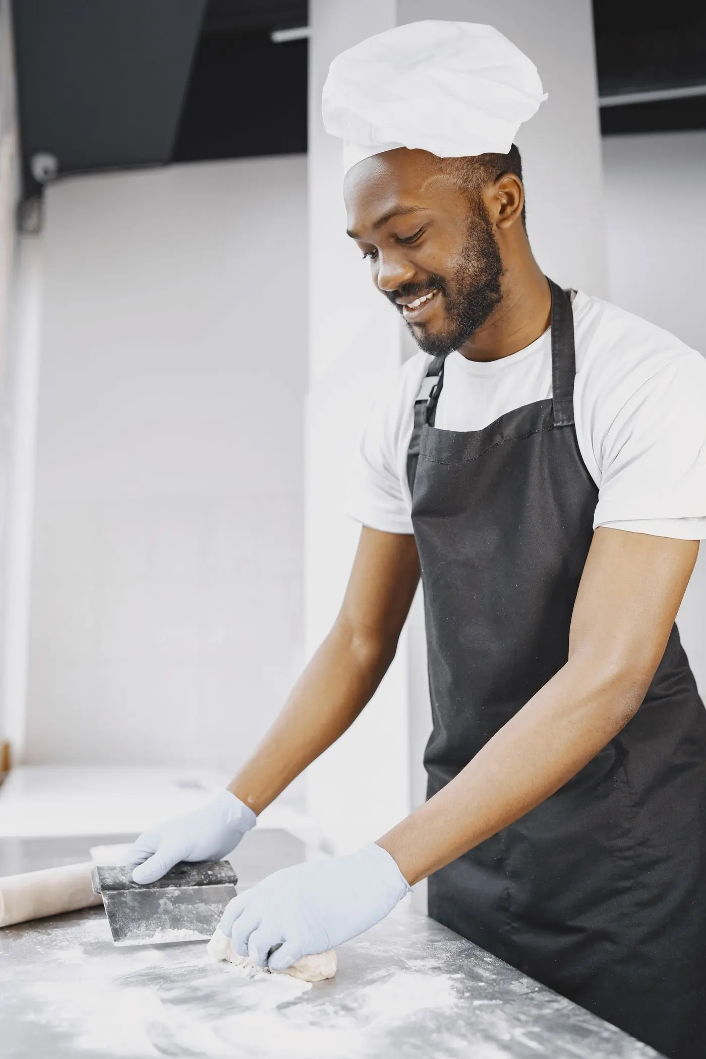A smiling baker in a white chef hat rolling dough in a bright room