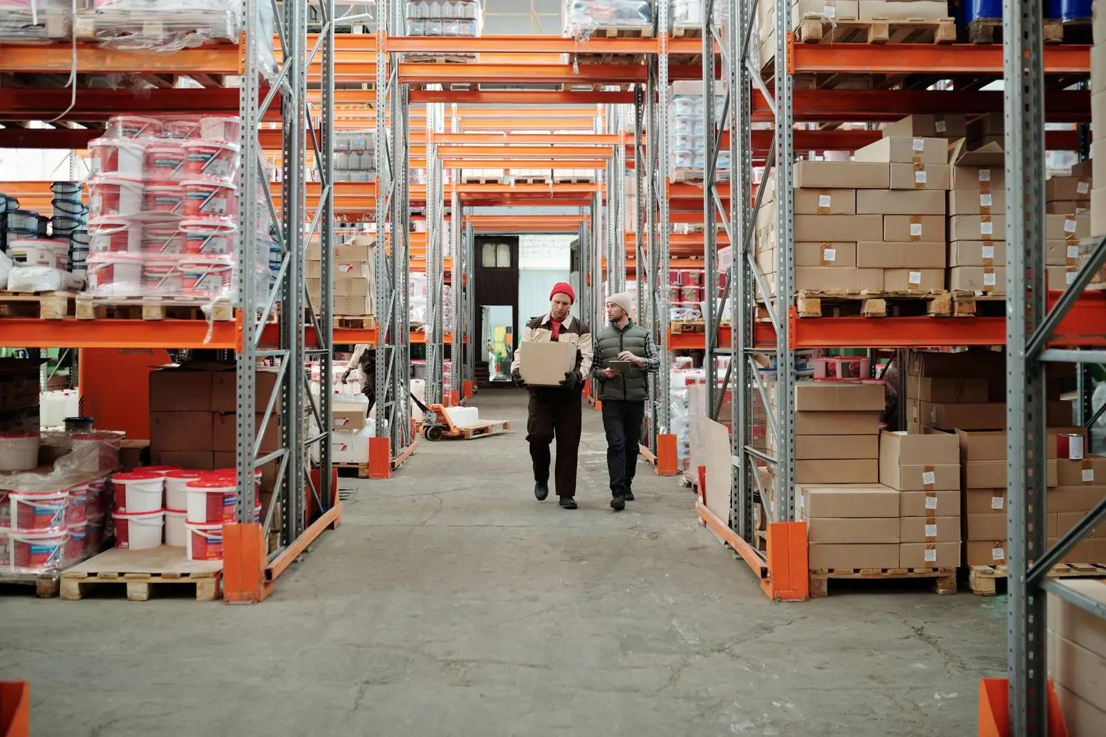 Two workers carrying a box down a tall orange warehouse aisle