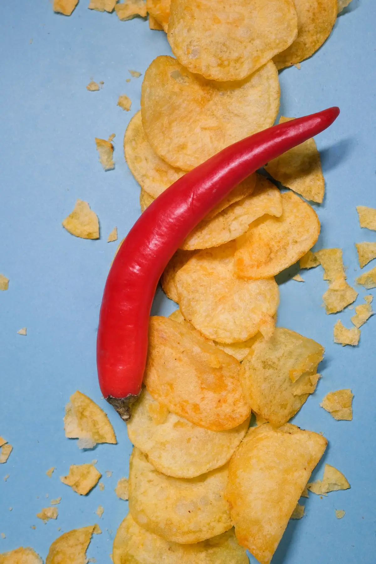 A red chilli pepper laid on a column of golden chips against powder blue