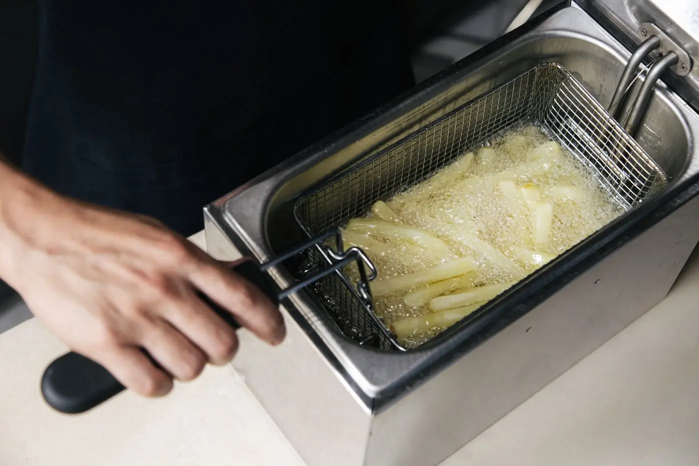 A hand steadying a small stainless kettle while potato sticks bubble in golden oil