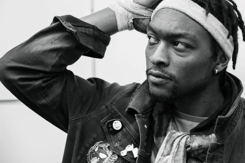 A man in a denim jacket with locs, photographed in black and white