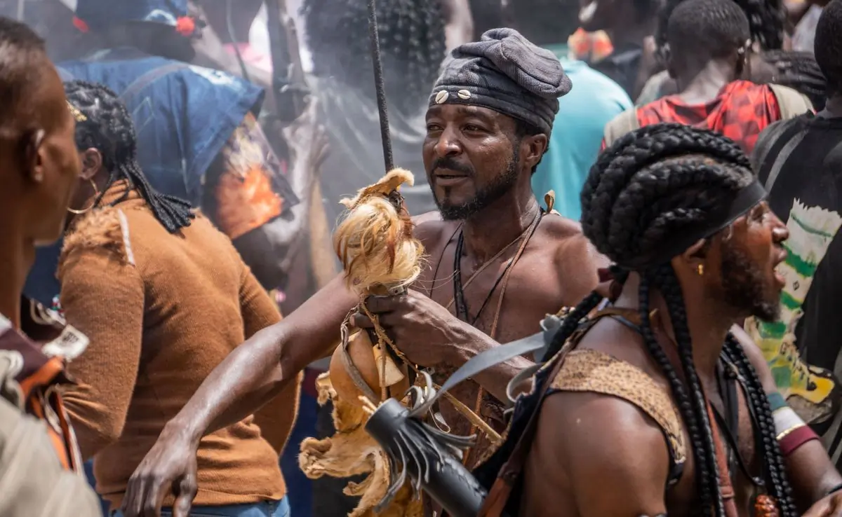A man with locs and beads at a street festival, surrounded by dancers